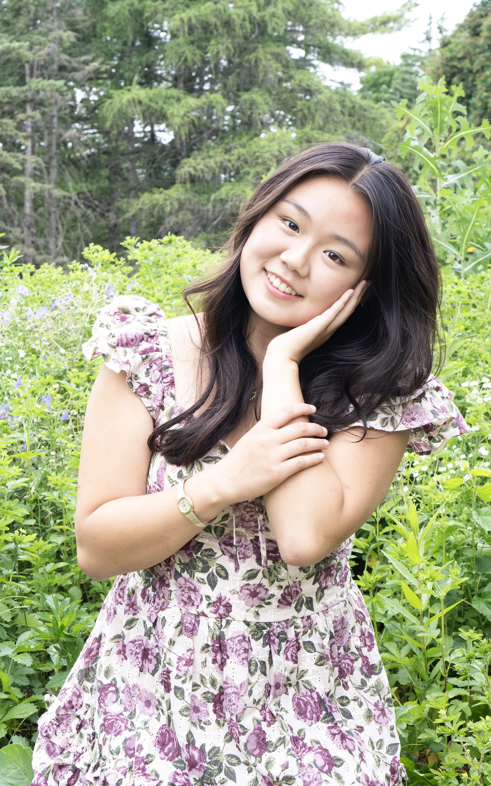 A young woman with long dark hair smiling in a floral dress in a lush green outdoor setting with trees and plants.