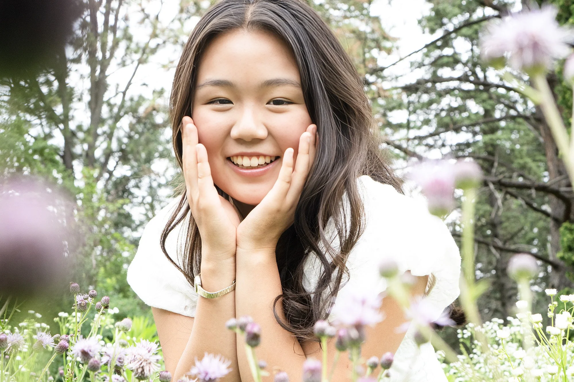 A smiling young woman with long dark hair and tan skin, holding her face with both hands, surrounded by a garden of pink and purple flowers with green foliage and trees in the background.