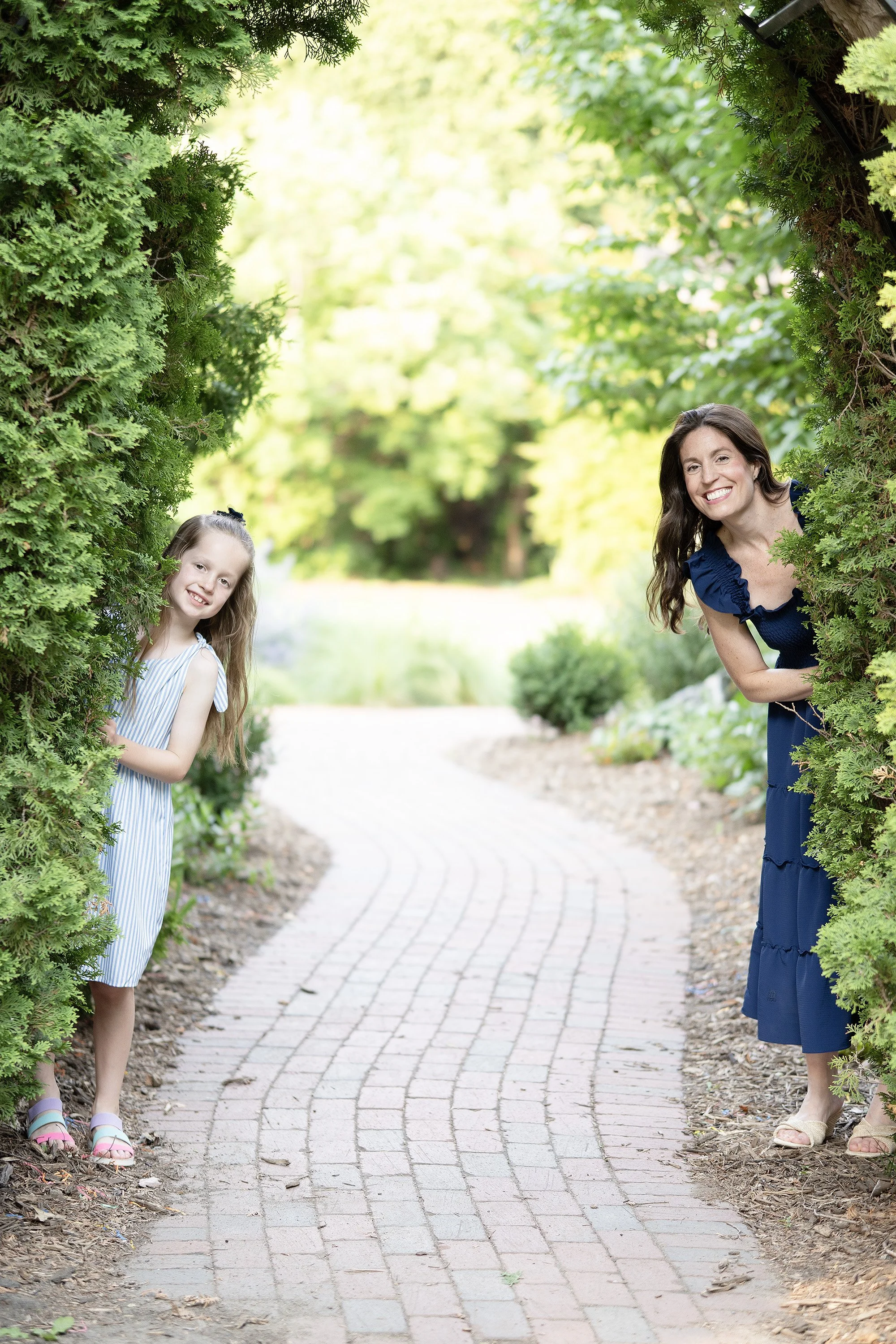 A woman and a girl peeking out from behind greenery on a curved brick pathway in a park.