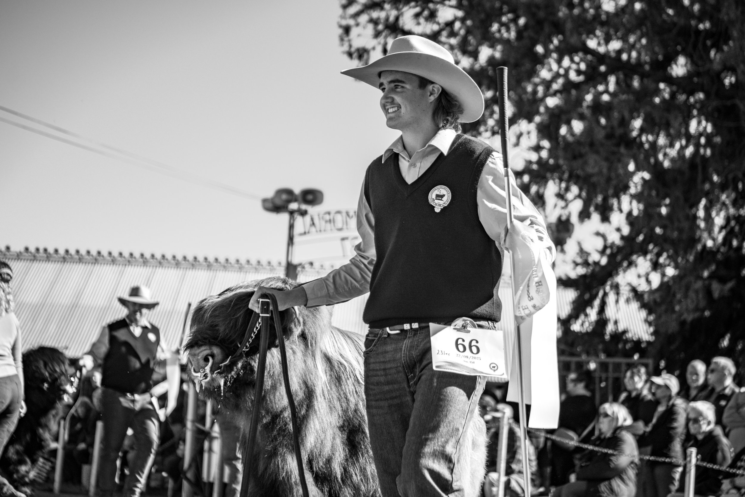 A young man in a cowboy hat and vest smiles while riding a horse at an outdoor event, with other riders and spectators visible in the background.