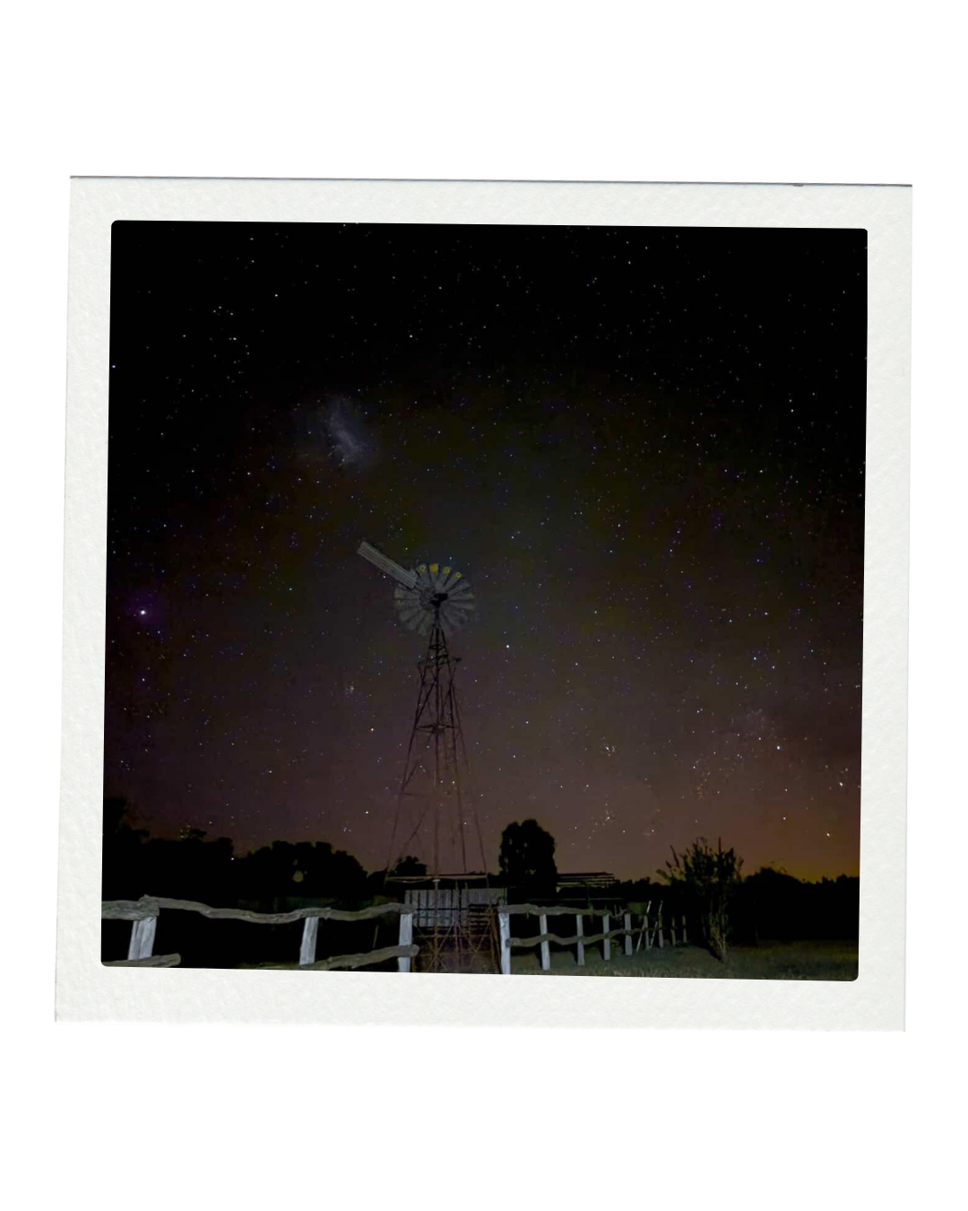 Night sky filled with stars, a windmill in the foreground, and a wooden fence below, captured during nighttime.