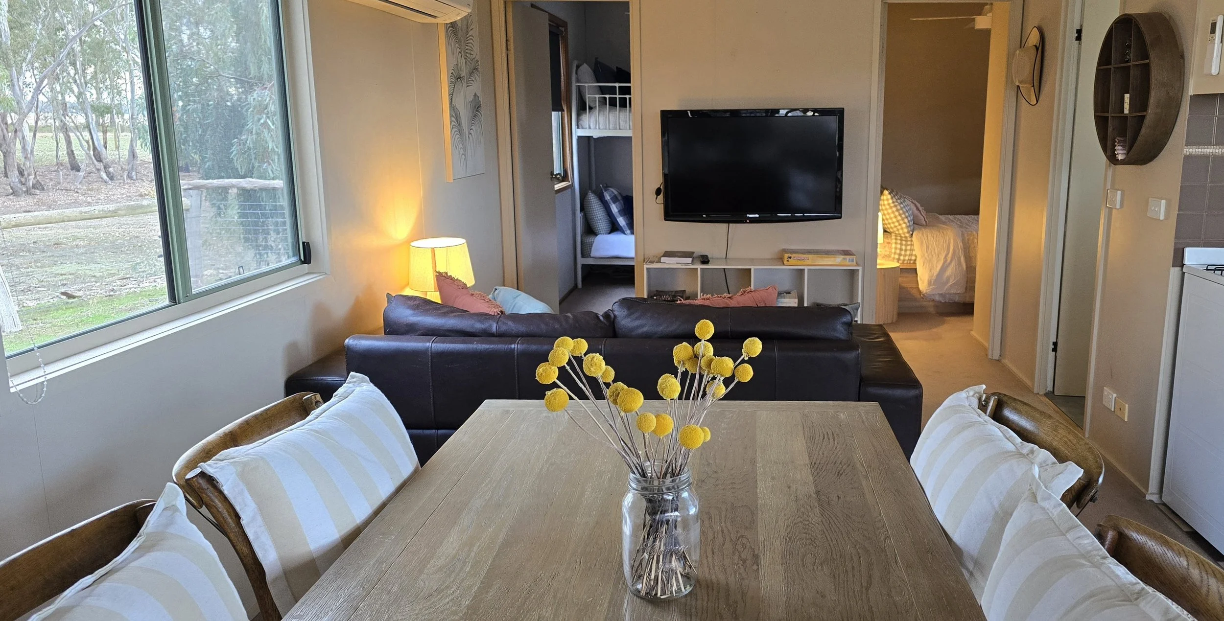 A cozy living room with a wooden dining table and six chairs, decorated with a glass vase full of yellow pom-pom flowers. The room features a large window to the left, a couch with pink pillows, a wall-mounted TV, and a small kitchenette to the right. In the background, there are two separate bedrooms, one with twin beds and the other with a larger bed. The room has warm lighting and a view of trees outside.
