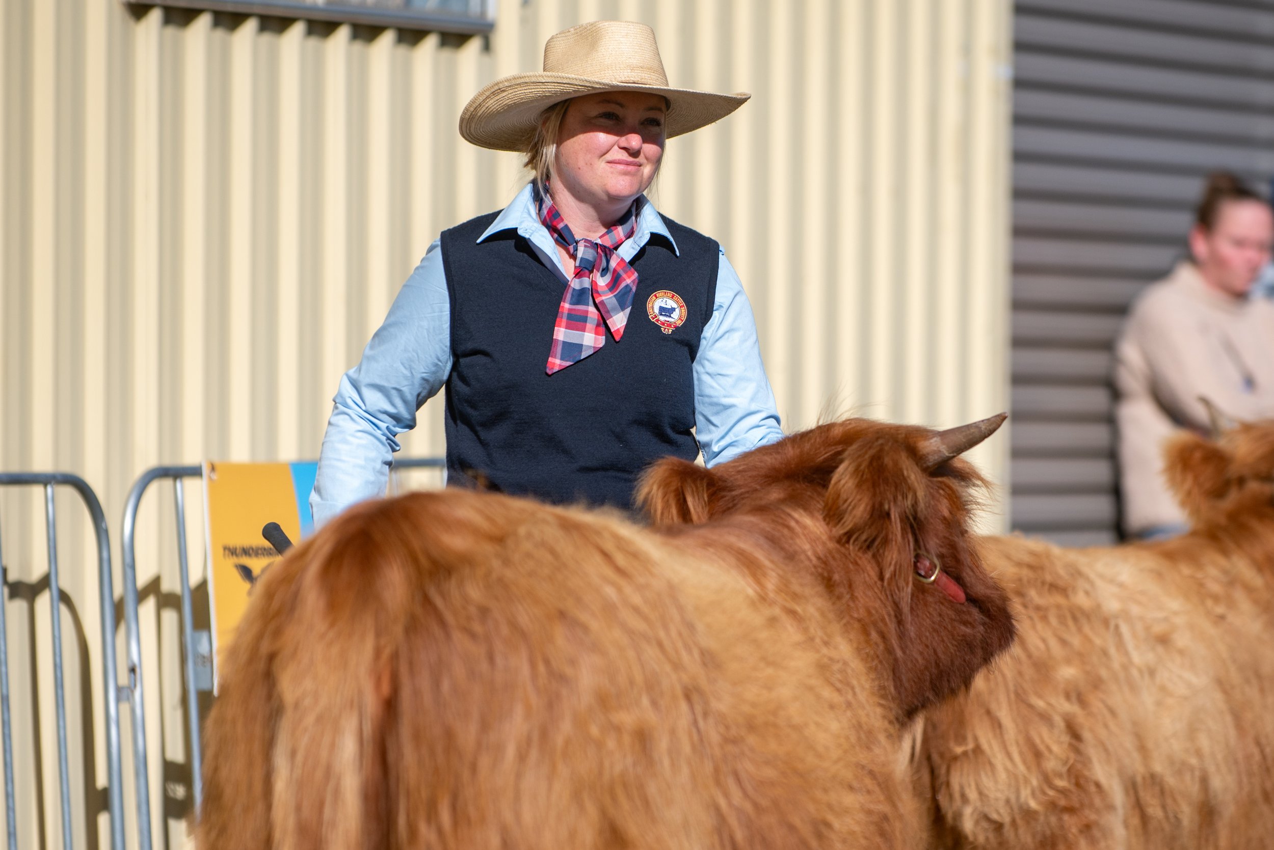 A woman wearing a large sun hat, blue shirt, and vest riding a brown horse at an outdoor event.