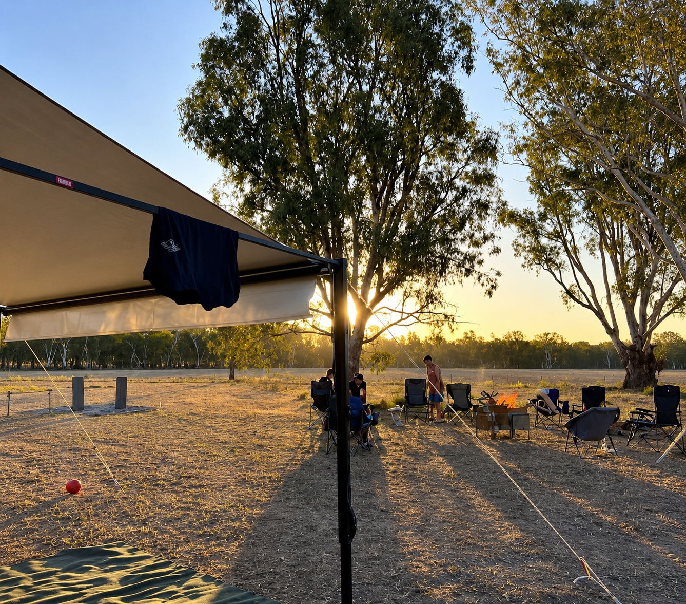 Group of people camping in an open field with folding chairs and a fire pit, under tall trees at sunset.