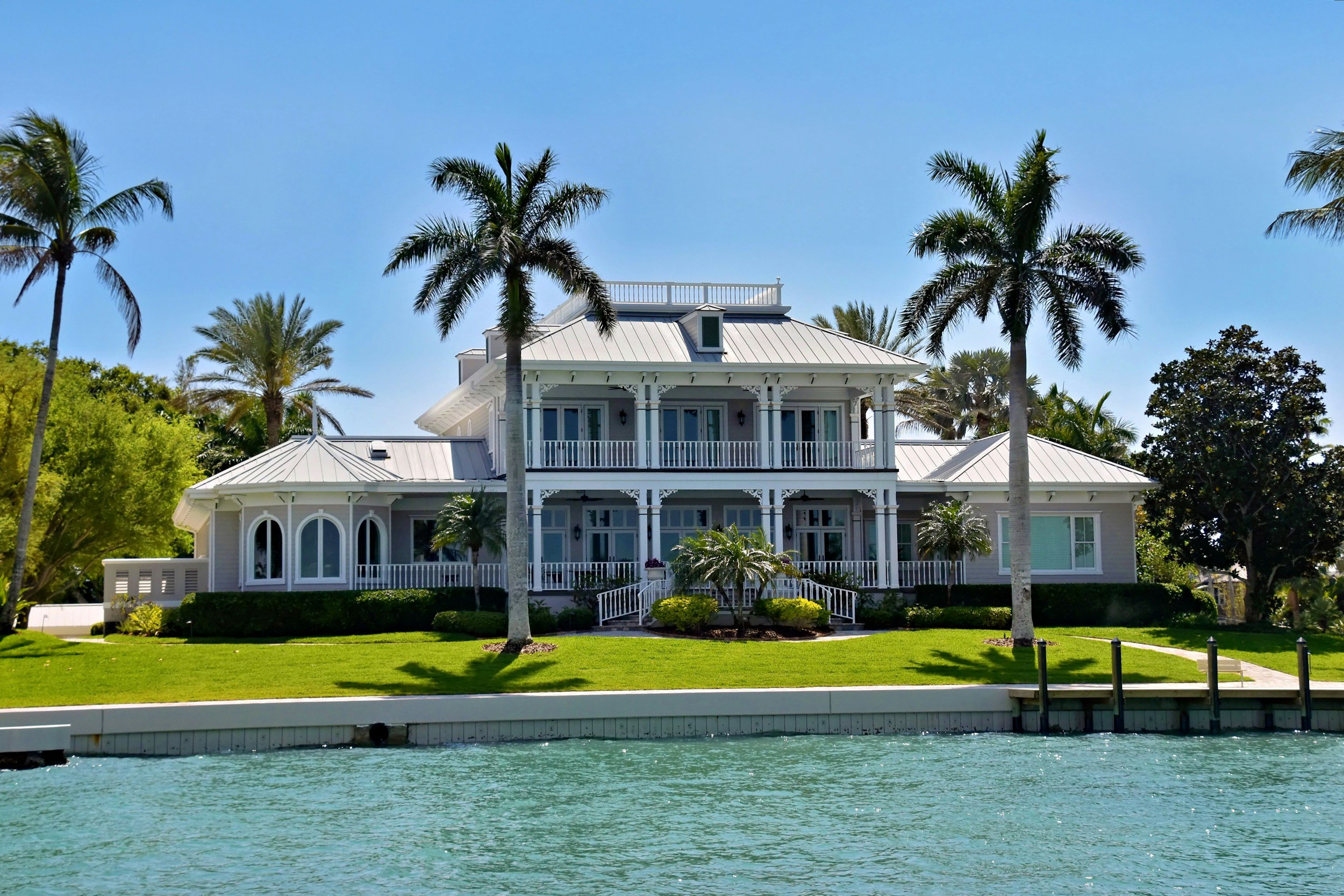 A large, white, Victorian-style house with a metal roof, multiple balconies, and garden landscaping, situated by water with palm trees around