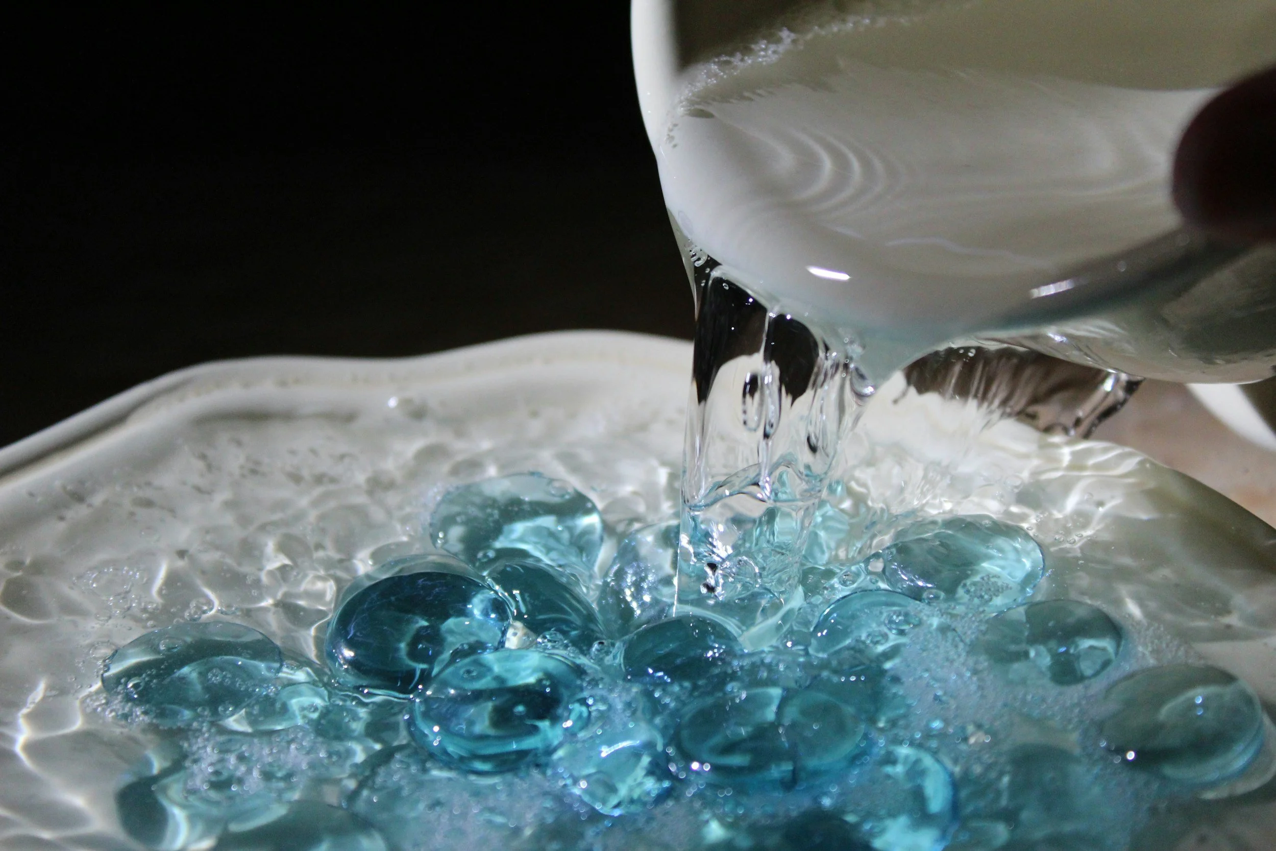 Pouring clear water into a bowl containing several small, blue glass marbles.