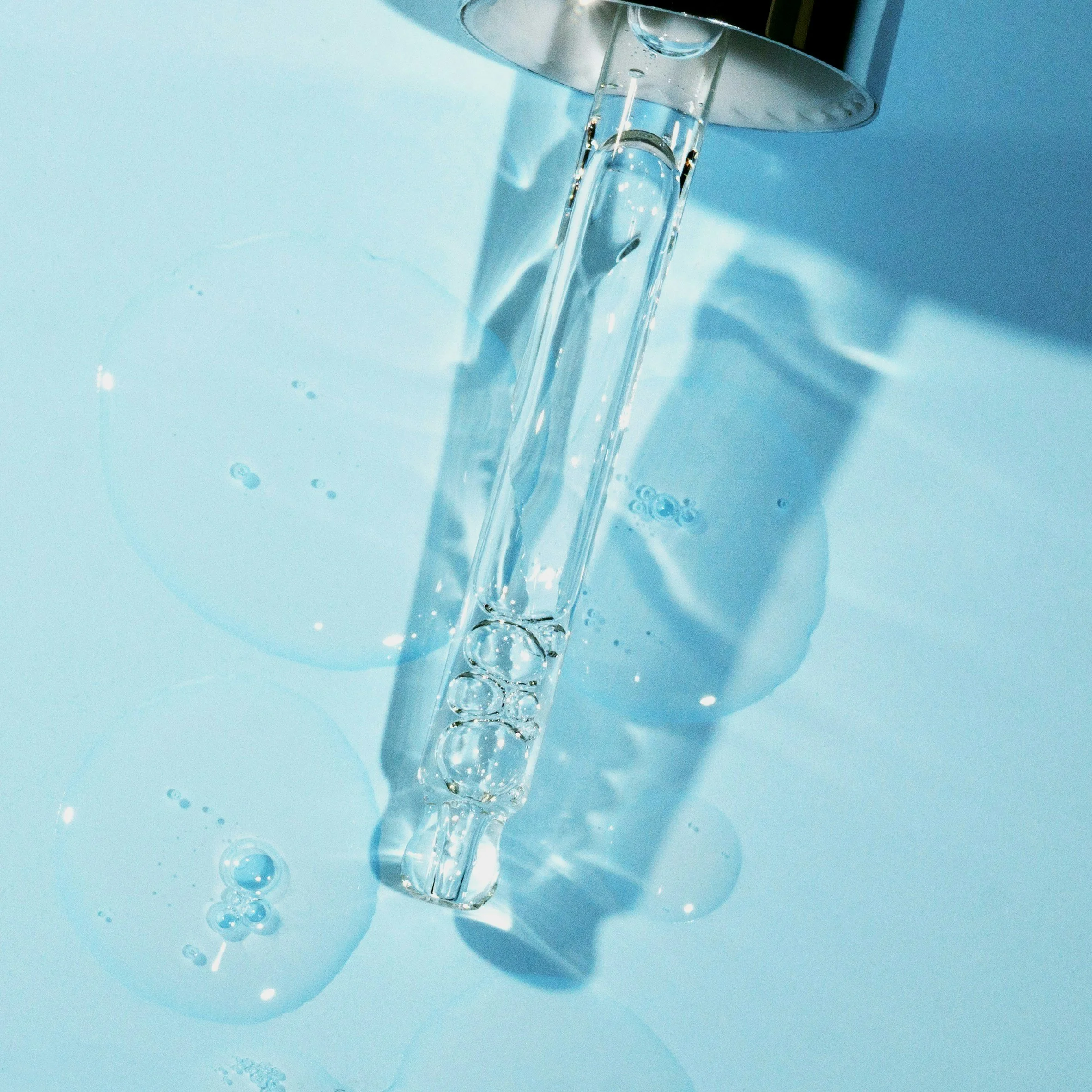 Close-up of a clear glass pipette with water inside, dripping into a surface, creating bubbles and ripples. The background is a light blue color.