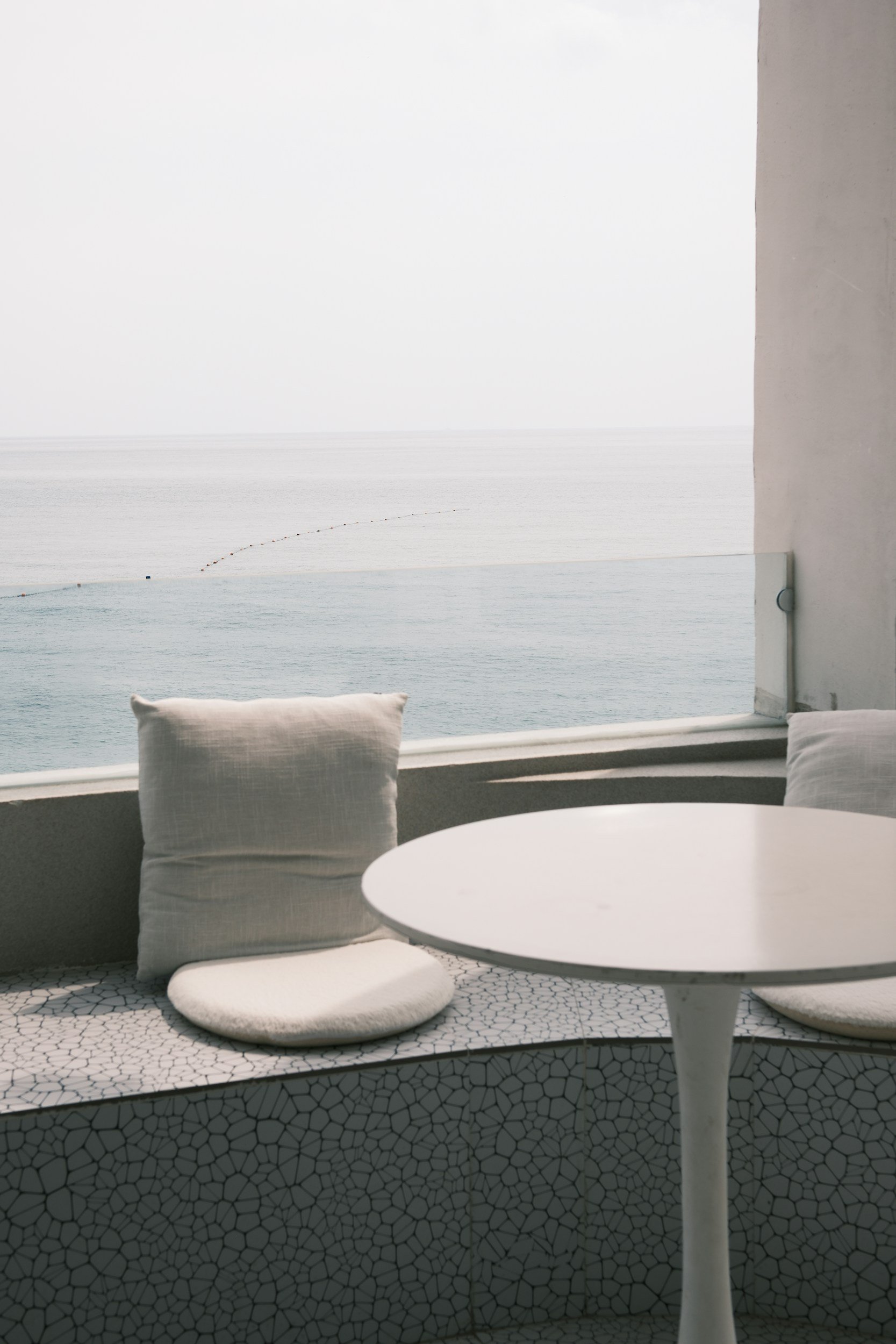 A white circular table with a white cushion on a bench with a black mosaic pattern, facing a large window with a view of the ocean and a white wall.