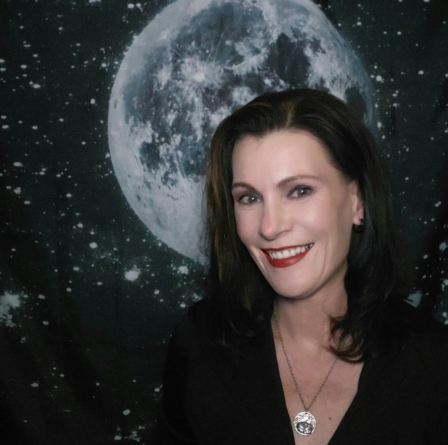 A woman with brown hair and a bright smile, wearing a black top and a necklace with a large circular pendant, standing in front of a backdrop featuring the moon and stars in space.