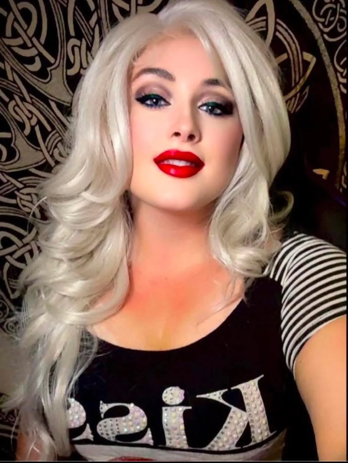 A woman with platinum blonde hair styled in loose curls, wearing bold red lipstick, dark eye makeup, and a black shirt with a rhinestone design on the front. She is lying against a decorative black and gold patterned background.