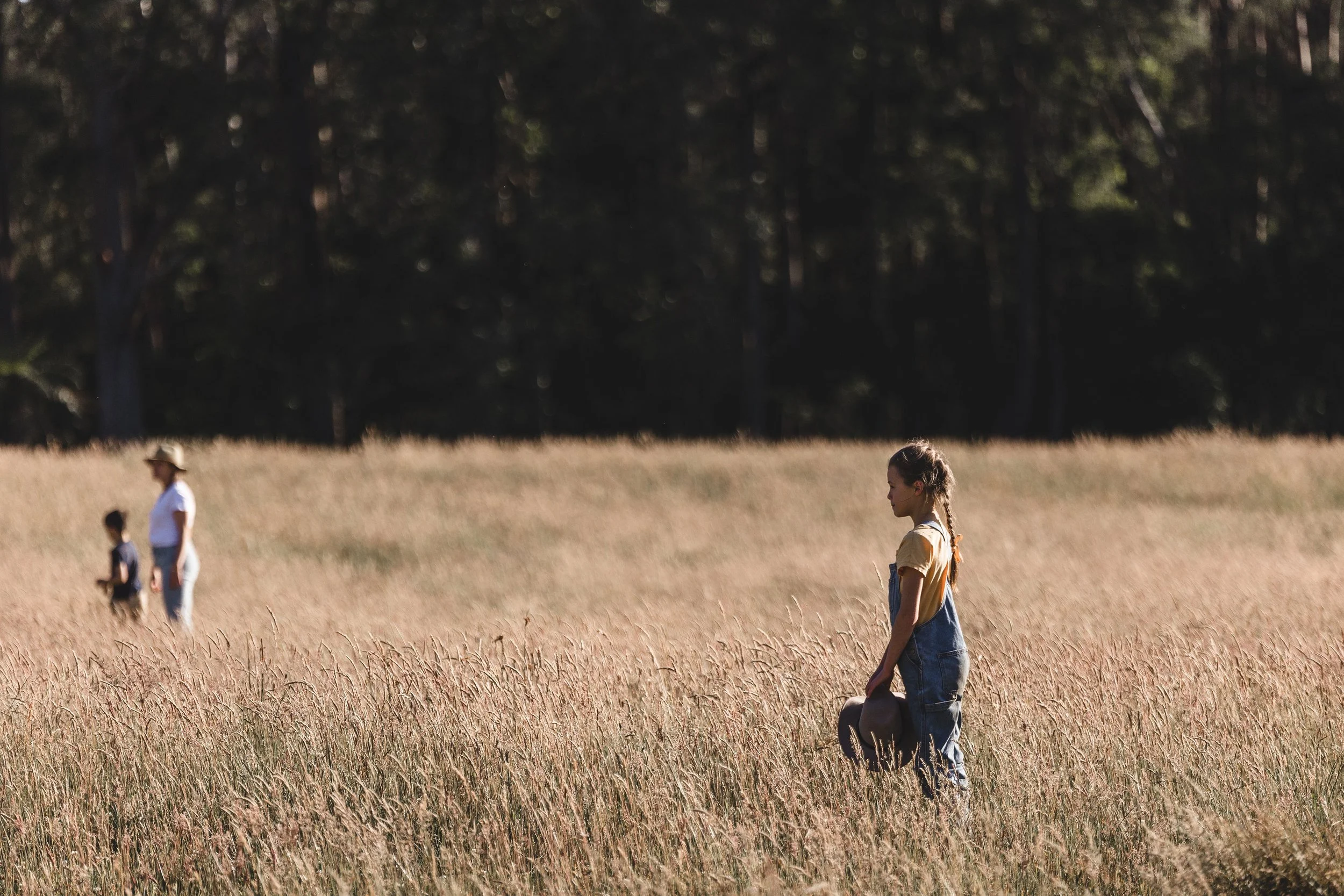 A girl walking through a field of tall, golden grass with trees in the background, other people can be seen in the distance.