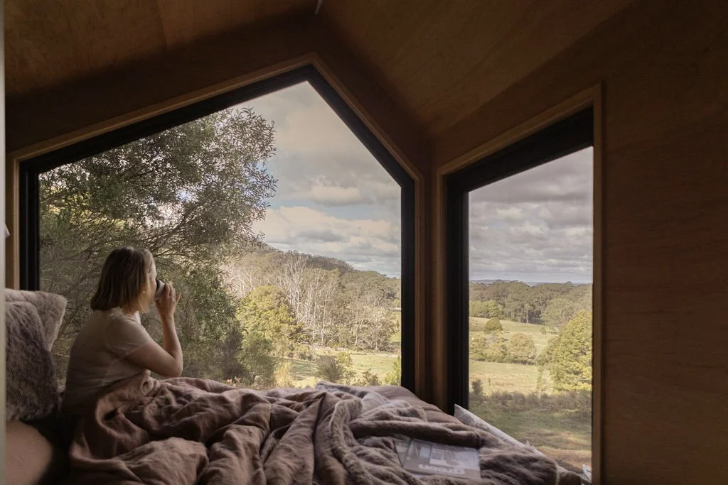 A person with short hair is sitting on a bed covered with a blanket, looking out a large window at the countryside with trees and cloudy sky.