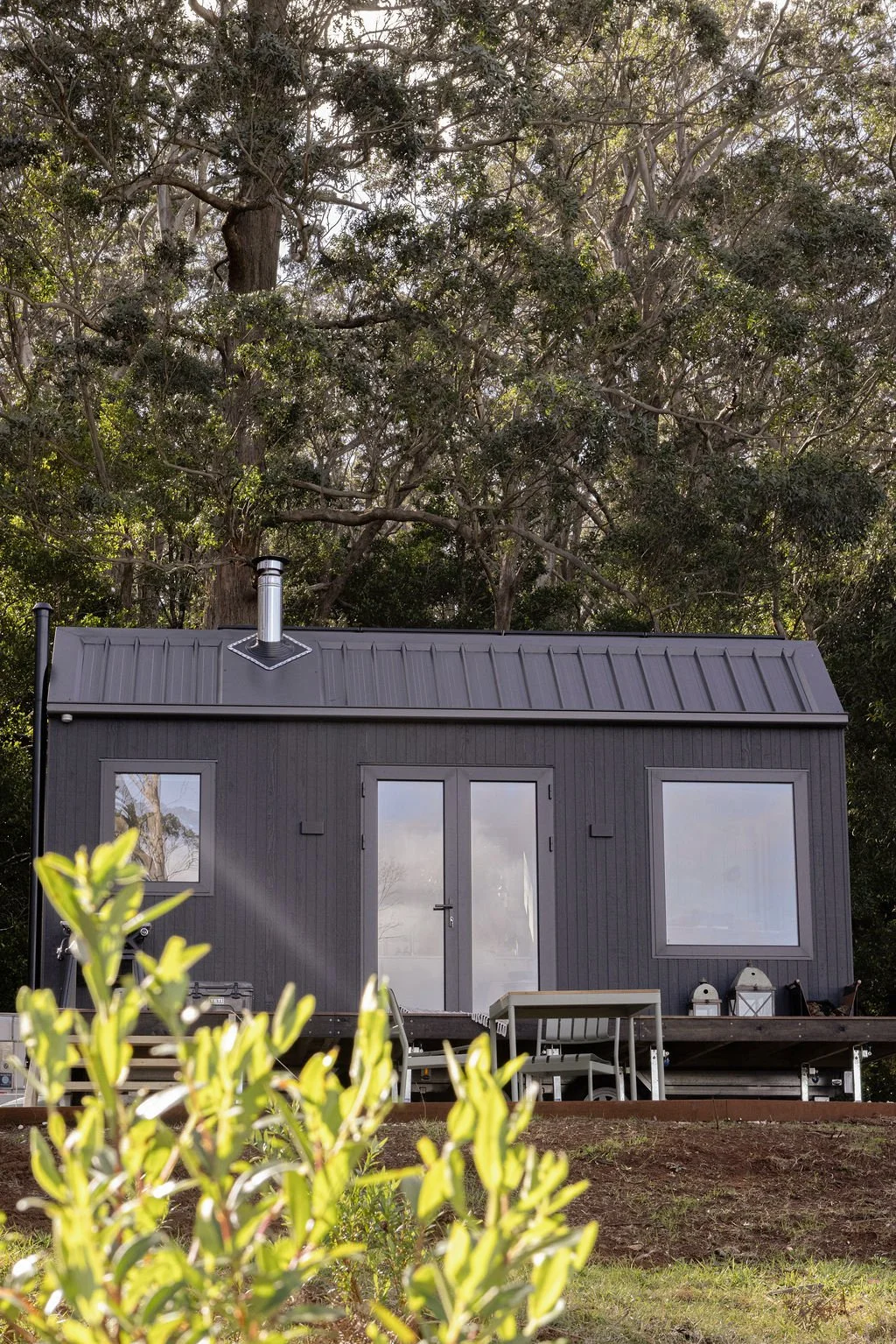 A small dark gray house with a metal roof, surrounded by trees. The house has a central door, two windows, and outdoor furniture and decorative items in front.