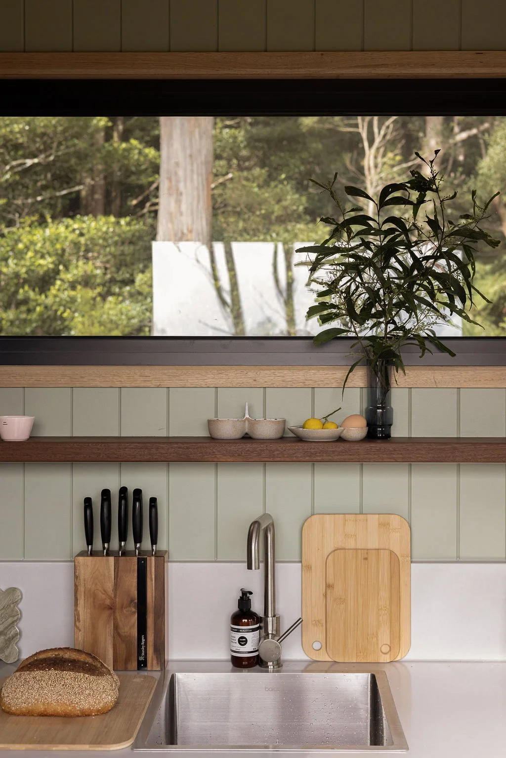 Kitchen window view showing trees and greenery outside with a potted plant inside on the countertop, kitchen utensils, cutting board, loaf of bread, and kitchen sink.