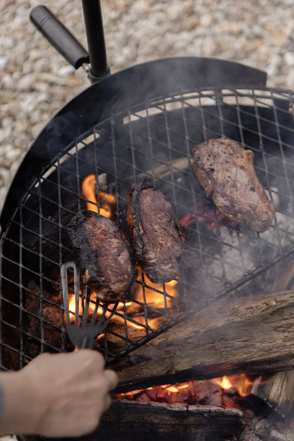 Three pieces of meat cooking on a grill over an open flame, with one hand holding a fork near the meat.