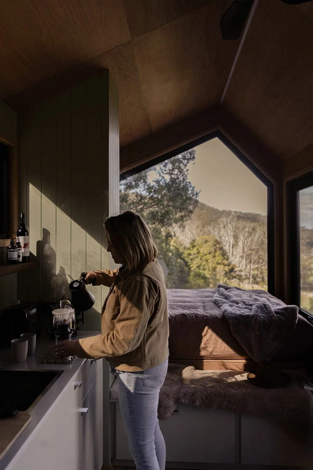 A woman pouring coffee in a cozy kitchen with a large window showing trees and mountains outside.