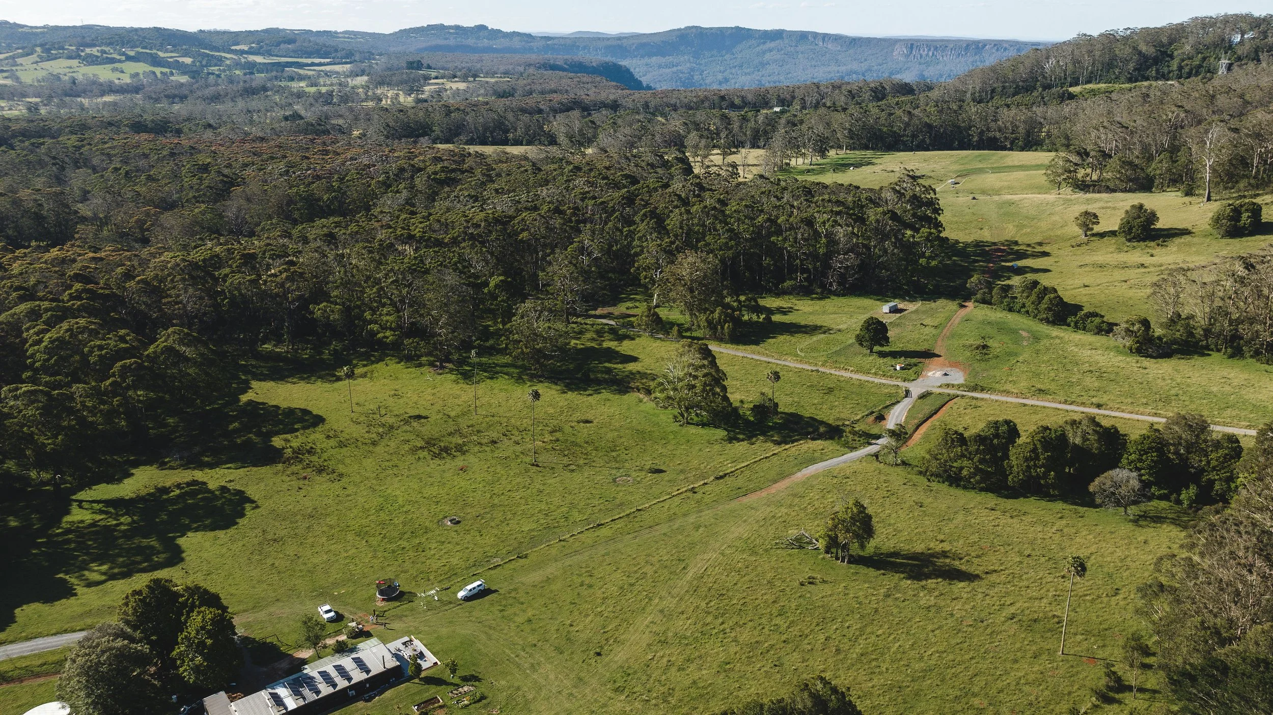 Aerial view of a countryside landscape with lush green fields, wooded areas, and a few scattered structures and vehicles. Dirt roads cross through the land, with rolling hills and distant mountains in the background.