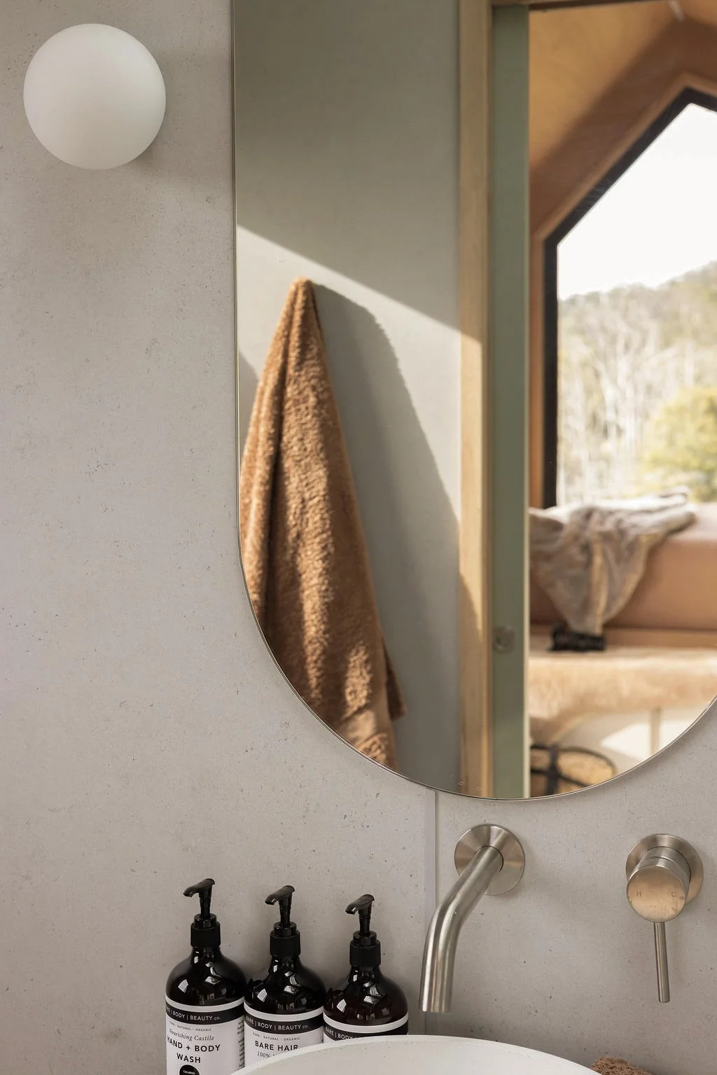 Close-up of a bathroom wall with a round mirror, three black bottles labeled hand and body wash, bare hair, and body lotion, a modern faucet, and a textured towel hanging on the wall. A window with a view of trees outside.