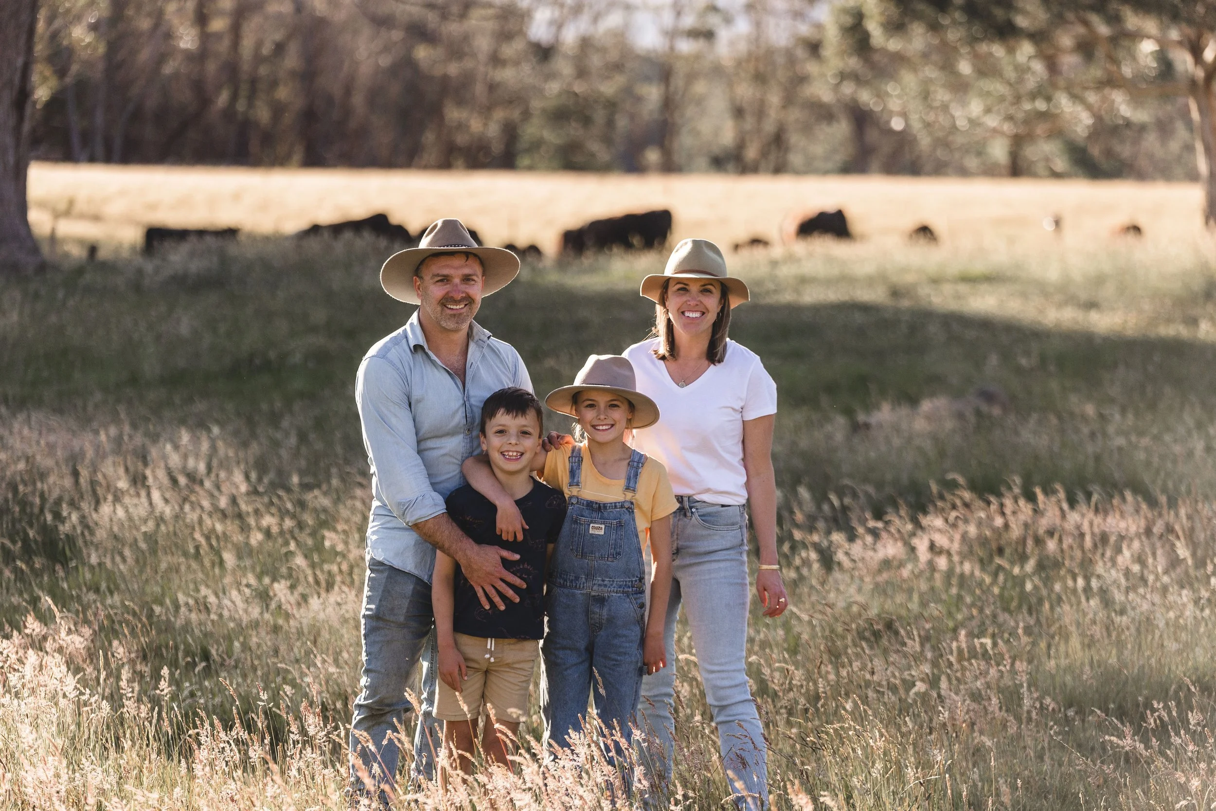 A happy family standing outdoors in a field of tall grass with trees in the background, all wearing hats and smiling.