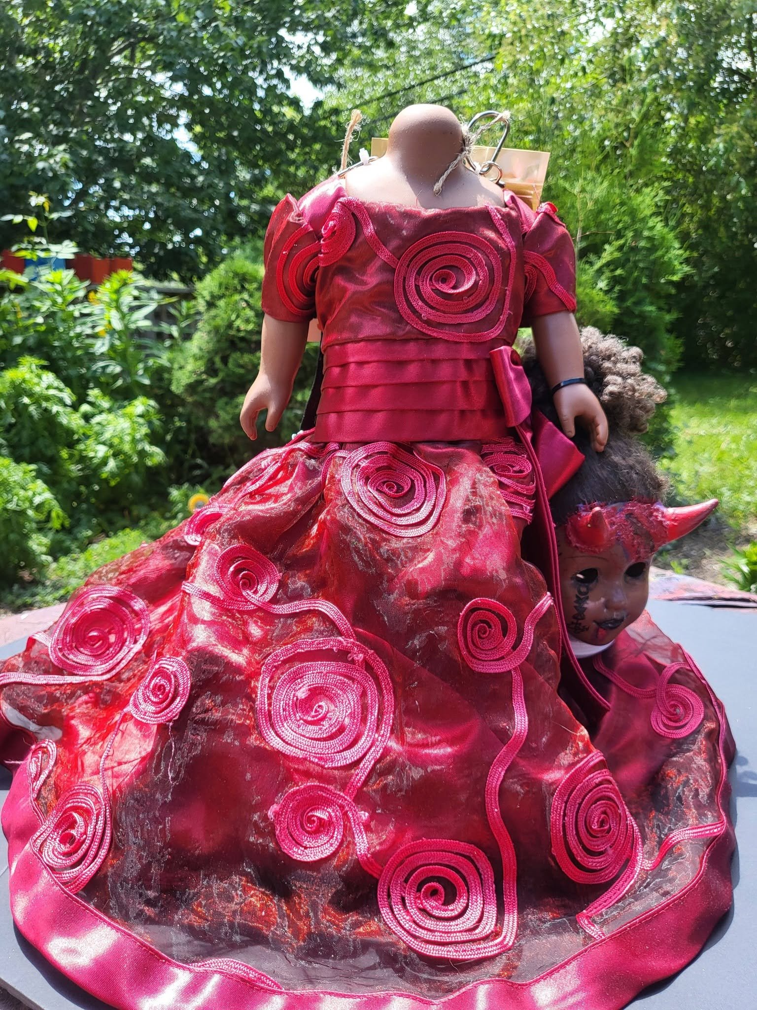 A doll dressed in a bright red, ruffled gown with swirl designs, standing outdoors against a backdrop of greenery.