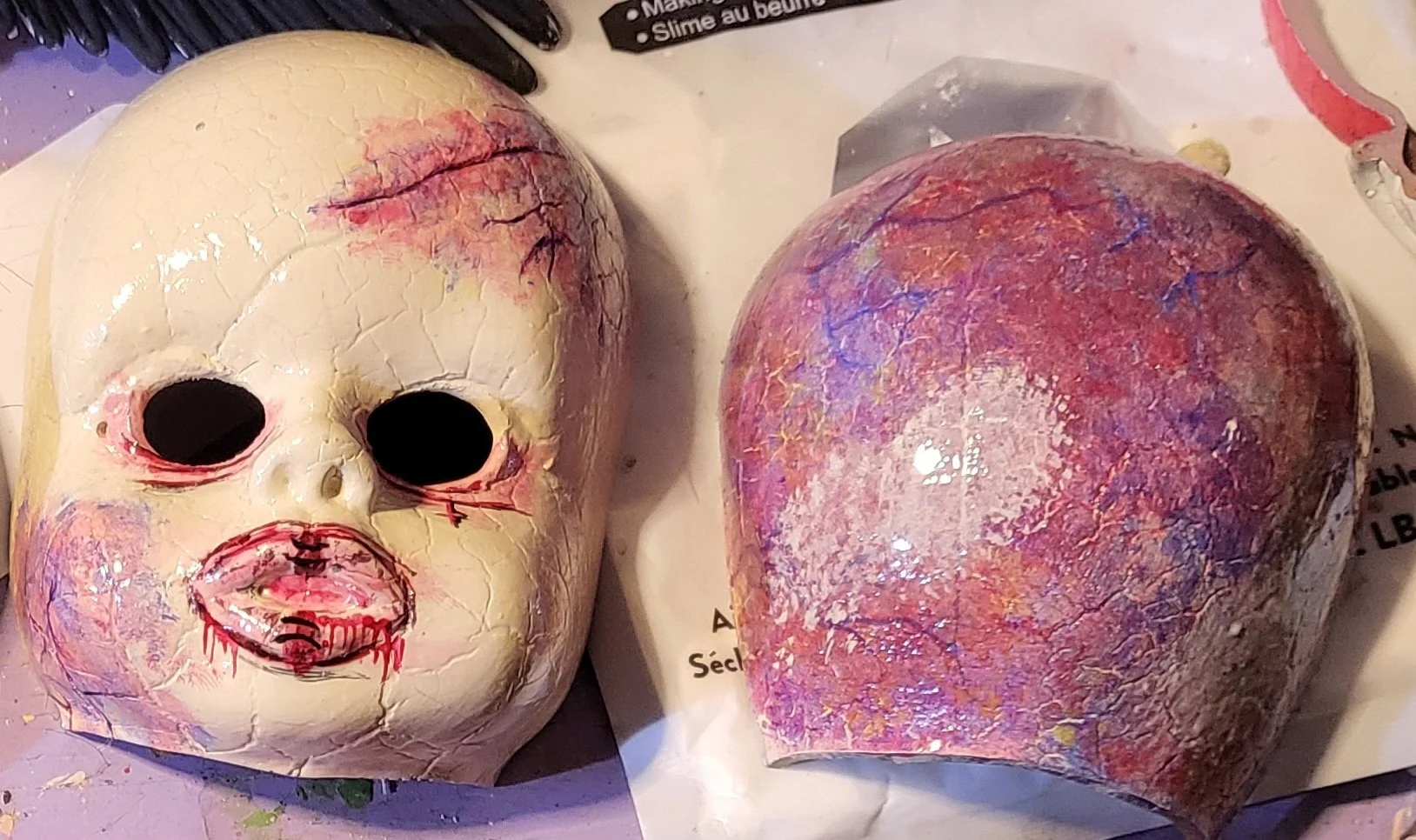 A broken Halloween mask resembling a monkey face with black eye sockets, pink painted lips, and cracks on the surface, next to a colorful heart-shaped object with purple, pink, and orange hues.