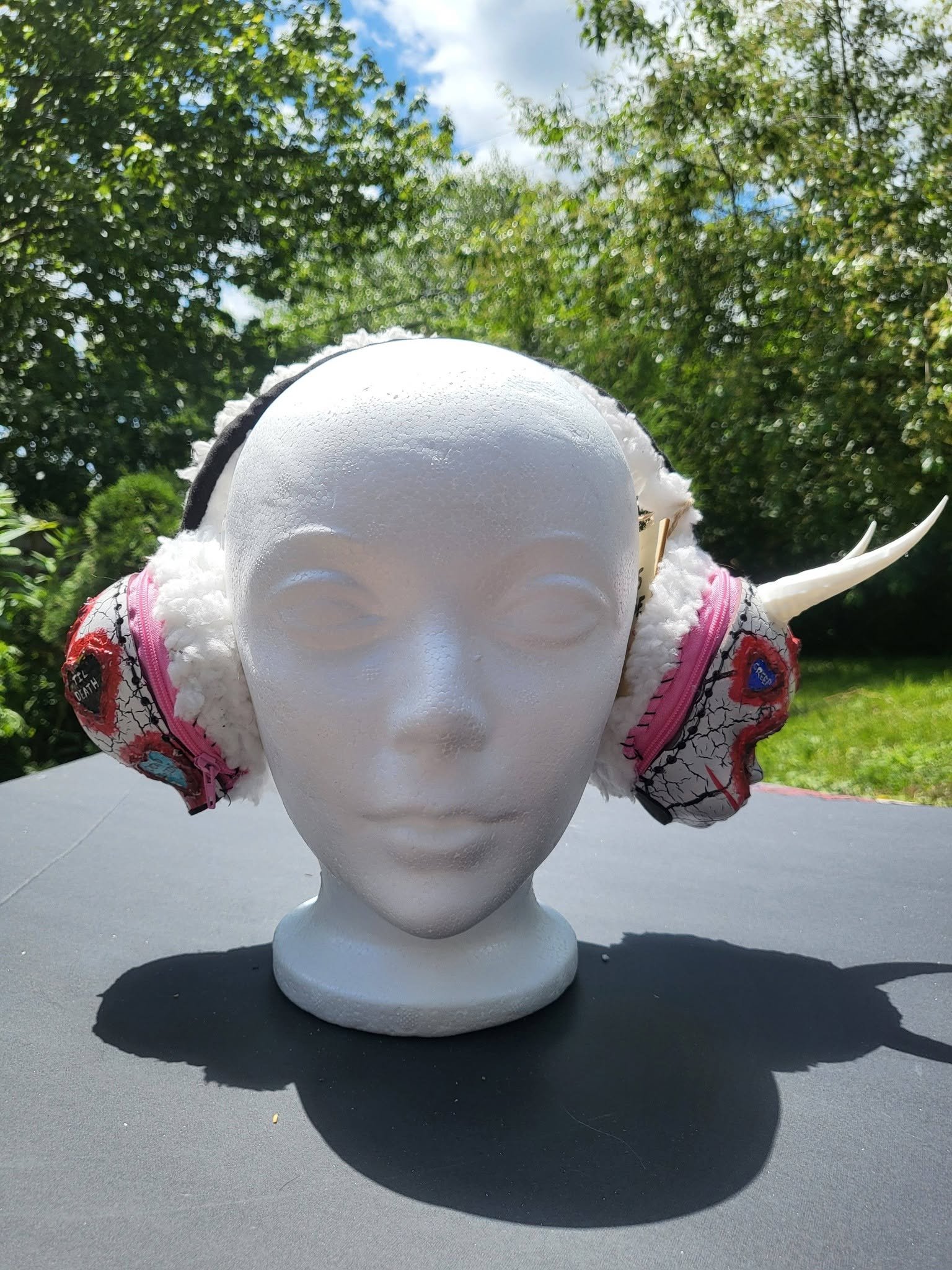 A white mannequin head wearing a pair of pink and black headphones with a zigzag pattern. The mannequin head is placed outdoors on a black surface with trees and a partly cloudy sky in the background.