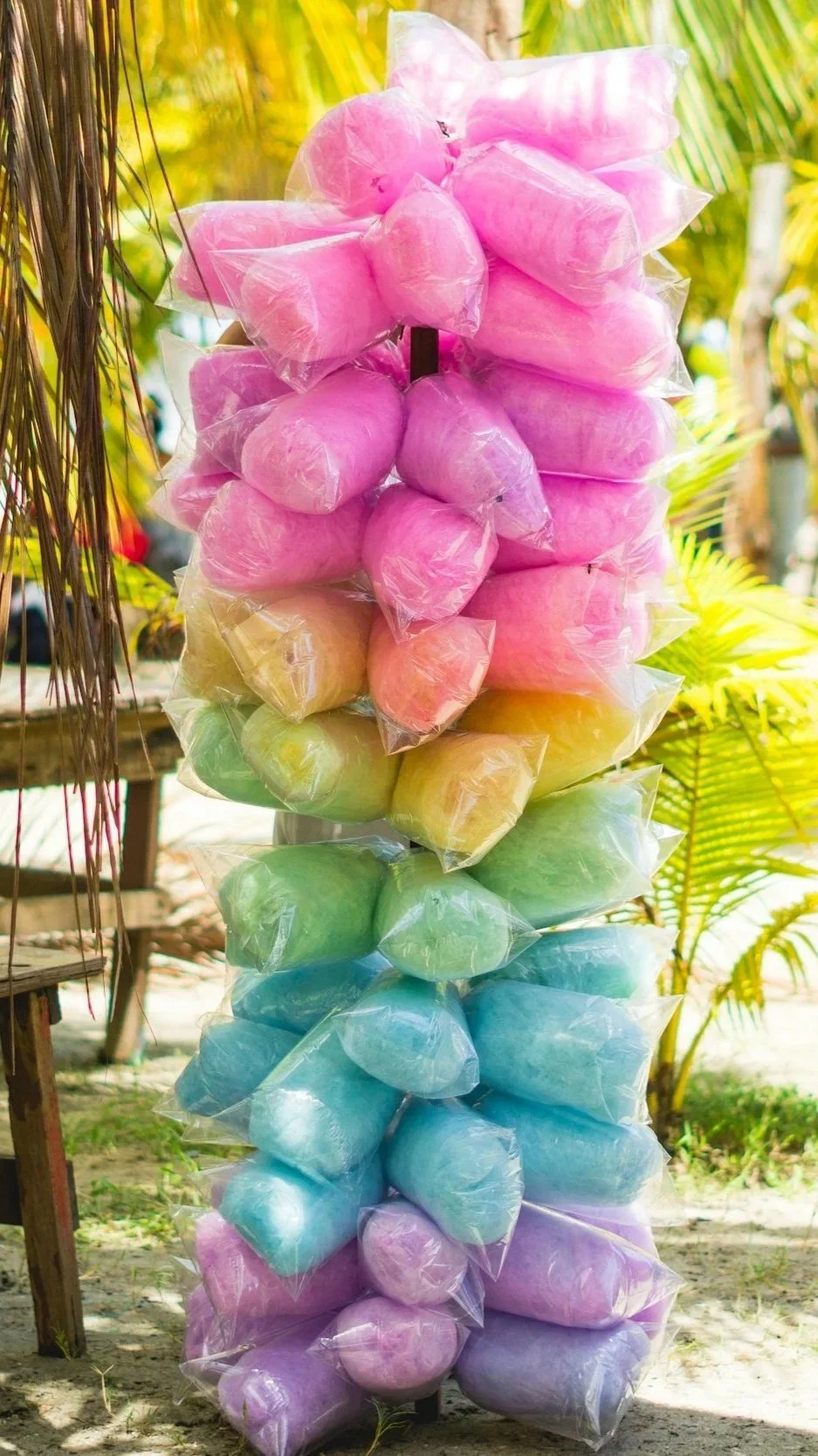 Colorful bags of cotton candy stacked on a pole, arranged in rainbow order from pink, yellow, green, blue, purple, and some white, set outdoors with palm tree leaves in the background.