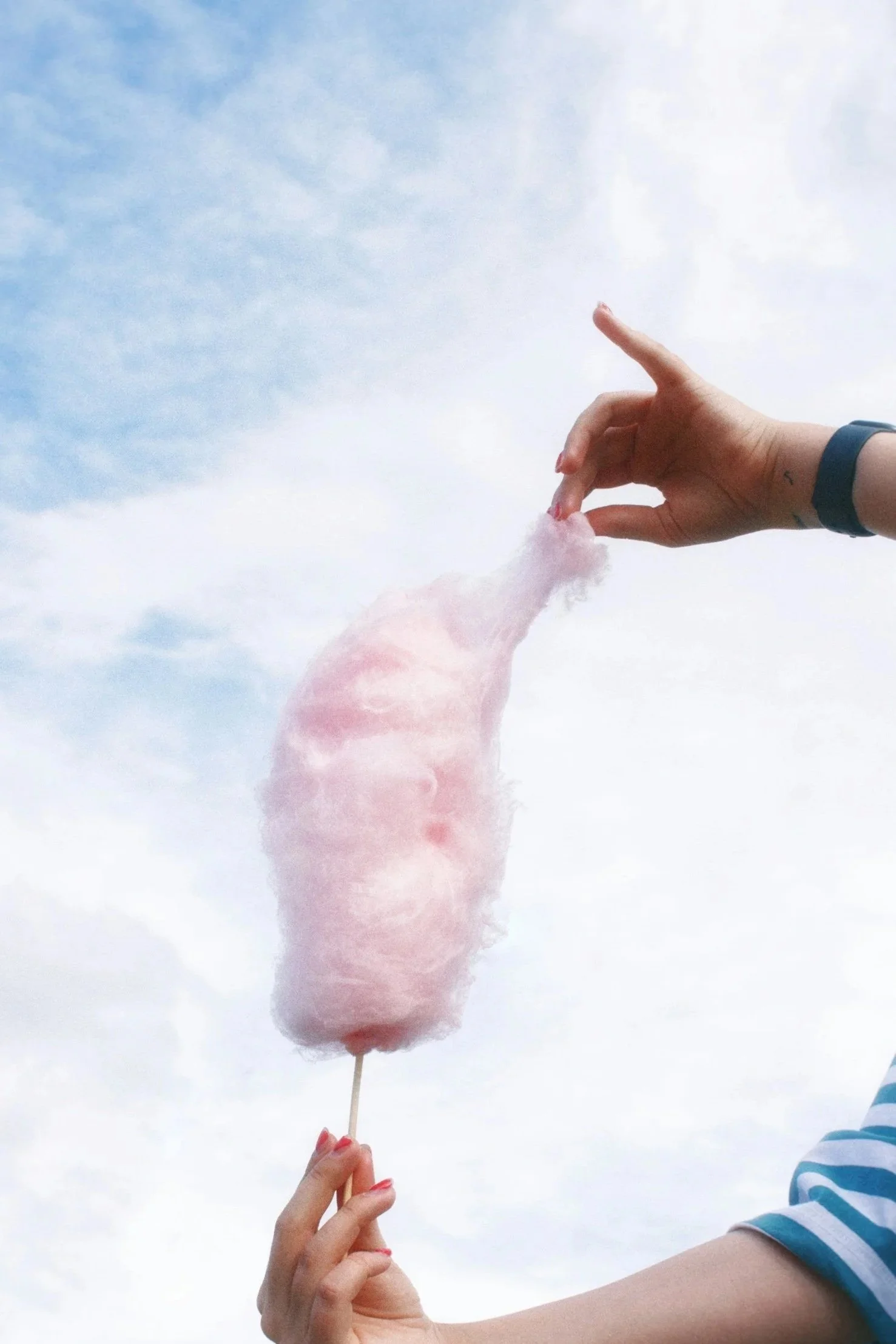Person holding pink cotton candy on a stick, with another hand holding the stick, against a cloudy sky.