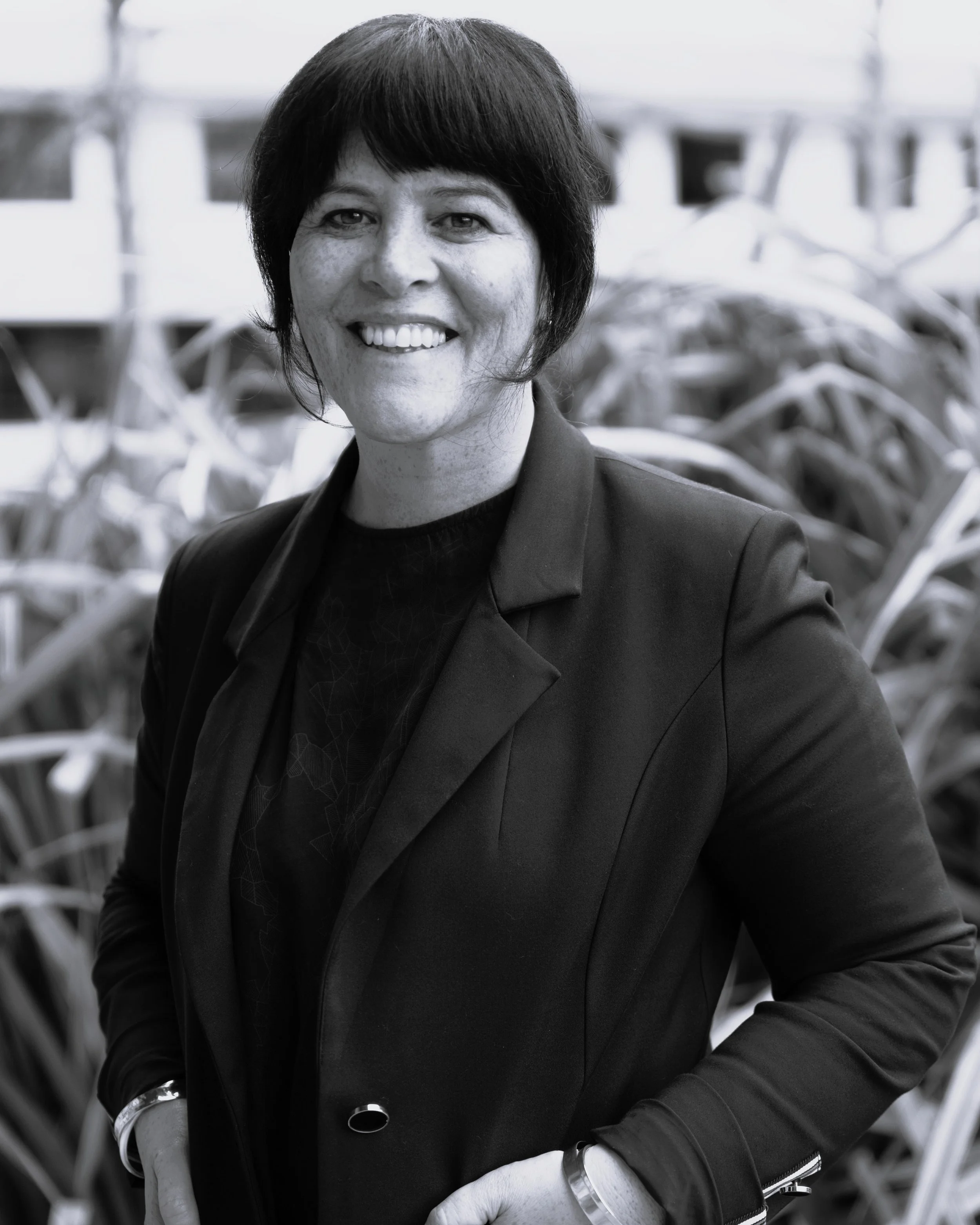 Black and white photo of a woman with short dark hair smiling, standing outdoors with blurred large plants or shrubs in the background.