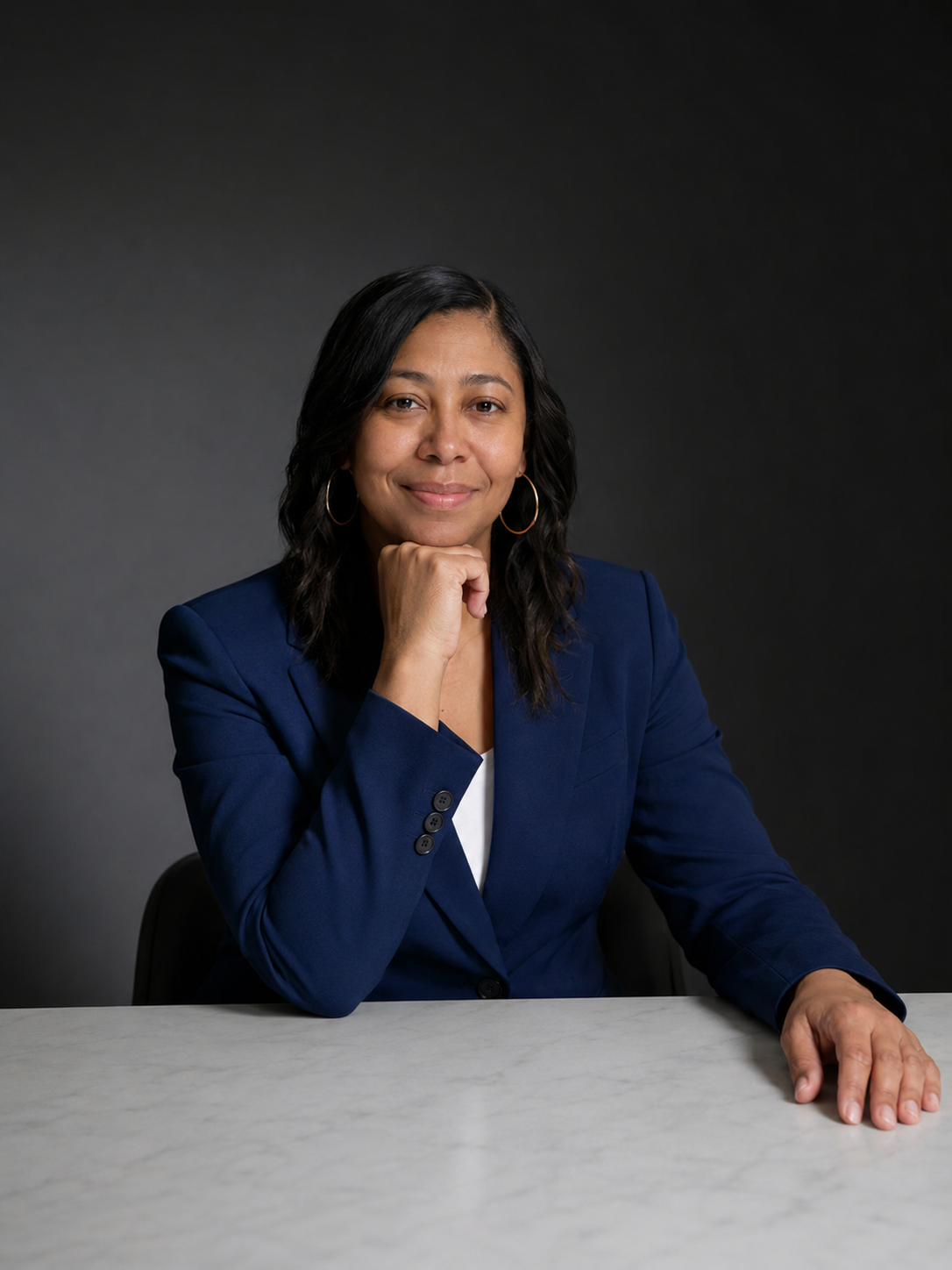A woman with black wavy hair wearing a navy blazer and hoop earrings, sitting at a marble table against a dark gray gradient background, resting her chin on her right hand and smiling.