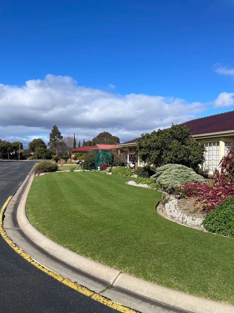 Residential neighborhood with well-maintained lawn, landscaped bushes, and houses with red roofs under a partly cloudy sky.