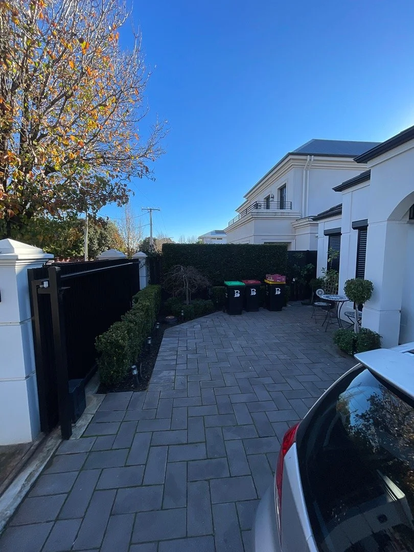 A paved driveway outside modern white houses with black accents, surrounded by trees and bushes, under a clear blue sky, with some trash bins and a parked car visible.