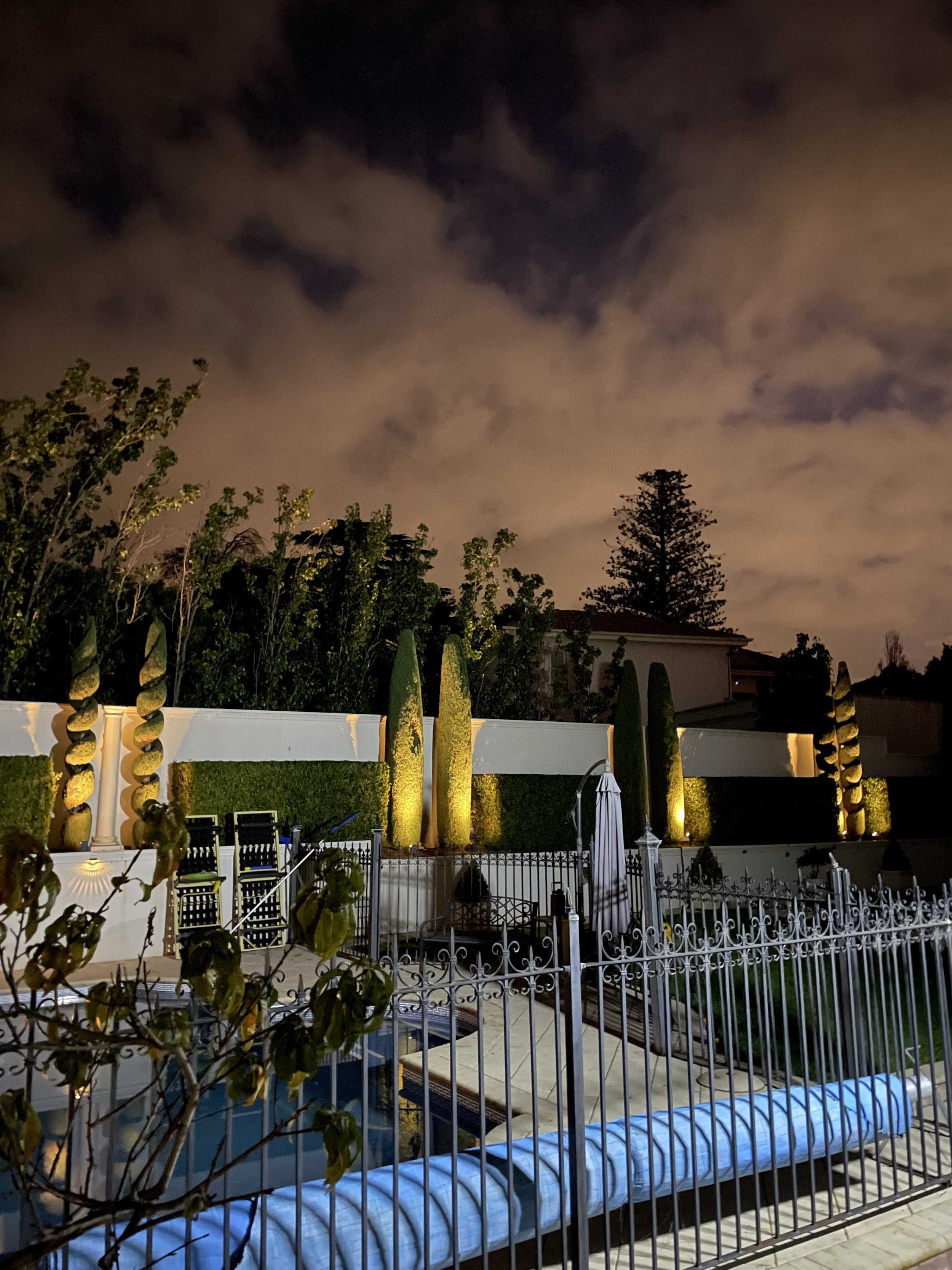 Nighttime backyard scene with a swimming pool, poolside lounge chairs with cushions, tall yellow-green manicured hedges, a white decorative fence, trees, a house roof, and a cloudy sky illuminated by ambient light.
