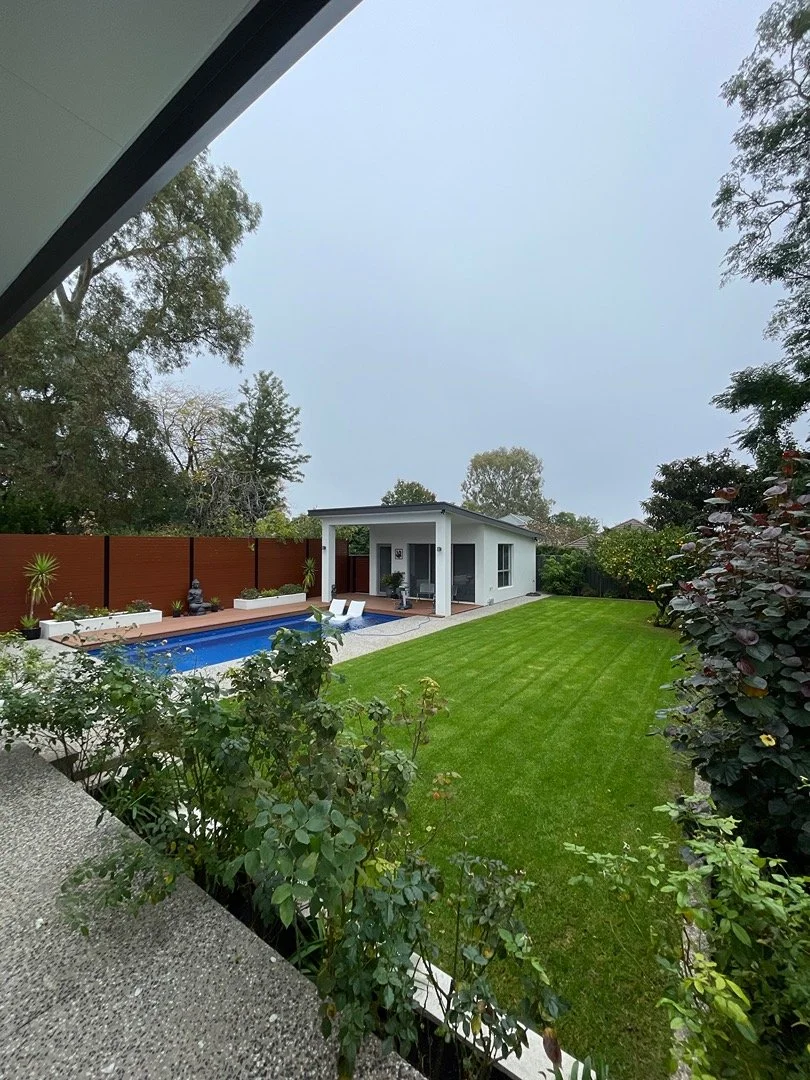 Backyard with a swimming pool, two lounge chairs, a small modern building, a wooden fence, lush green lawn, and trees in the background.