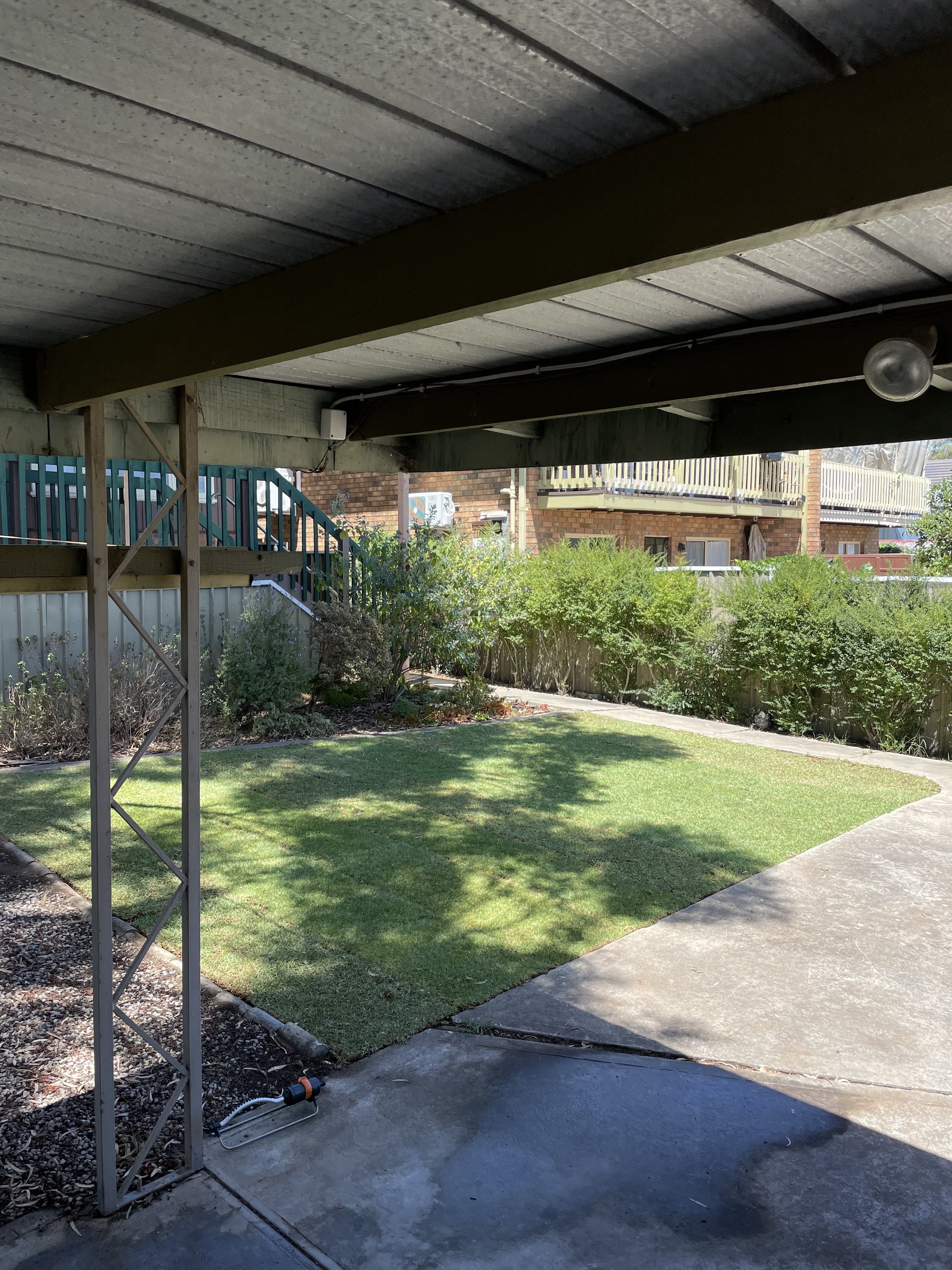 View of a backyard area with a well-maintained lawn, surrounding bushes, and a concrete patio. An upper level is visible, supported by wooden beams, with a house in the background that has a brick exterior and a balcony.