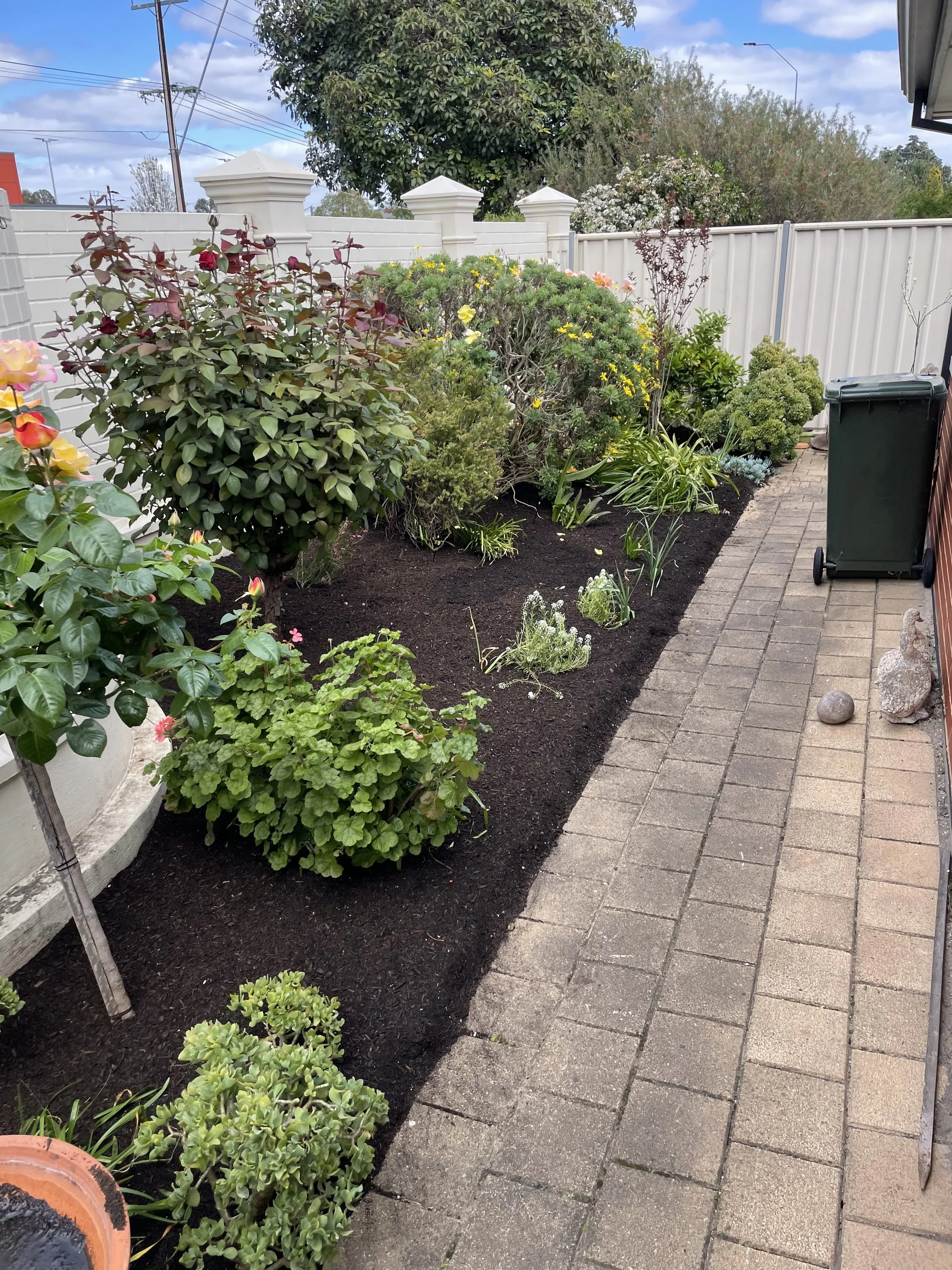 A backyard garden with flowering plants, shrubs, and a paved walkway bordered by freshly turned soil; white fence surrounds the yard under a partly cloudy sky.