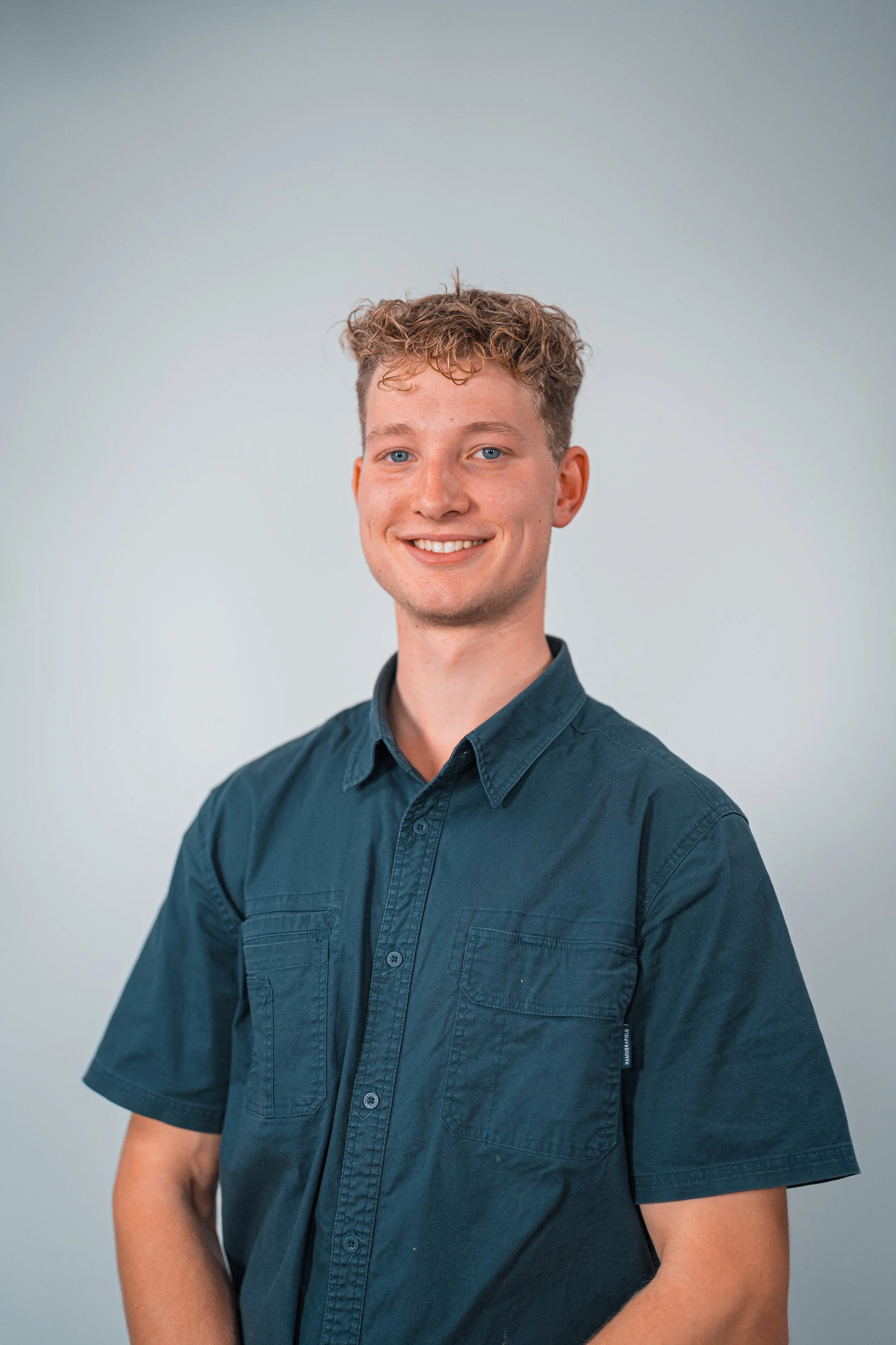 A young man with short, curly hair, blue eyes, and light skin, smiling and wearing a dark teal button-up shirt against a plain light grey background.