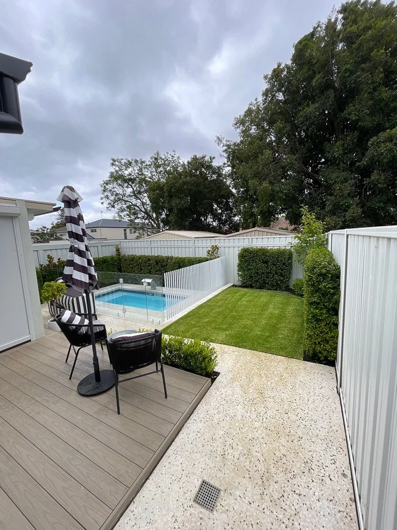 Backyard with a small swimming pool surrounded by a glass fence, a wooden deck with outdoor seating and an umbrella, a lawn, and tall trees with cloudy sky overhead.