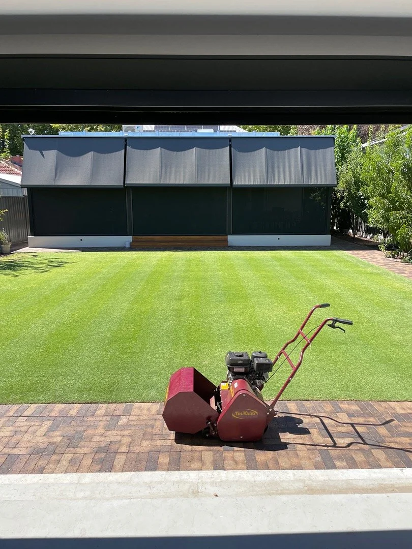 A red lawn roller on a brick patio, with a green lawn and a dark-colored shed in the background under a partly cloudy sky.