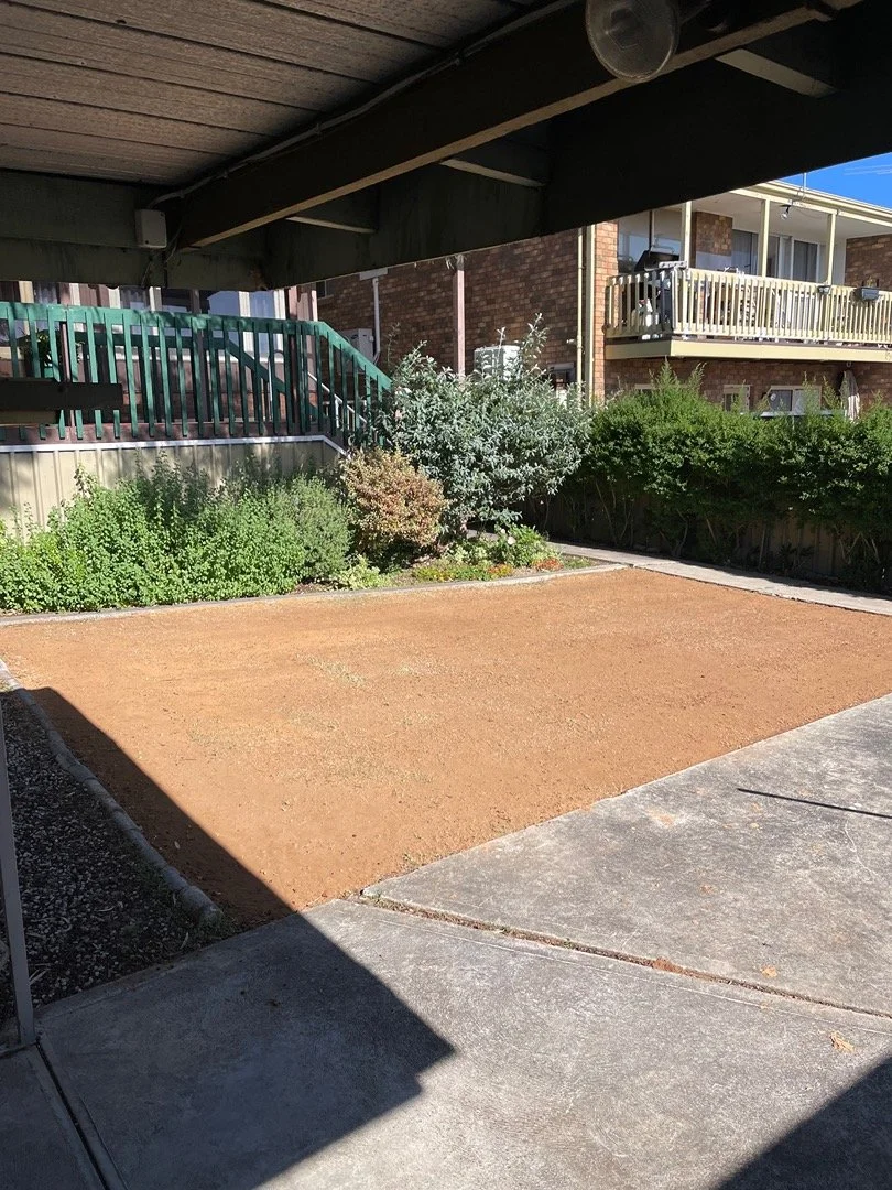Empty backyard with a dirt patch surrounded by some bushes and plants, with a porch overhead and neighboring apartment buildings with balconies in the background.