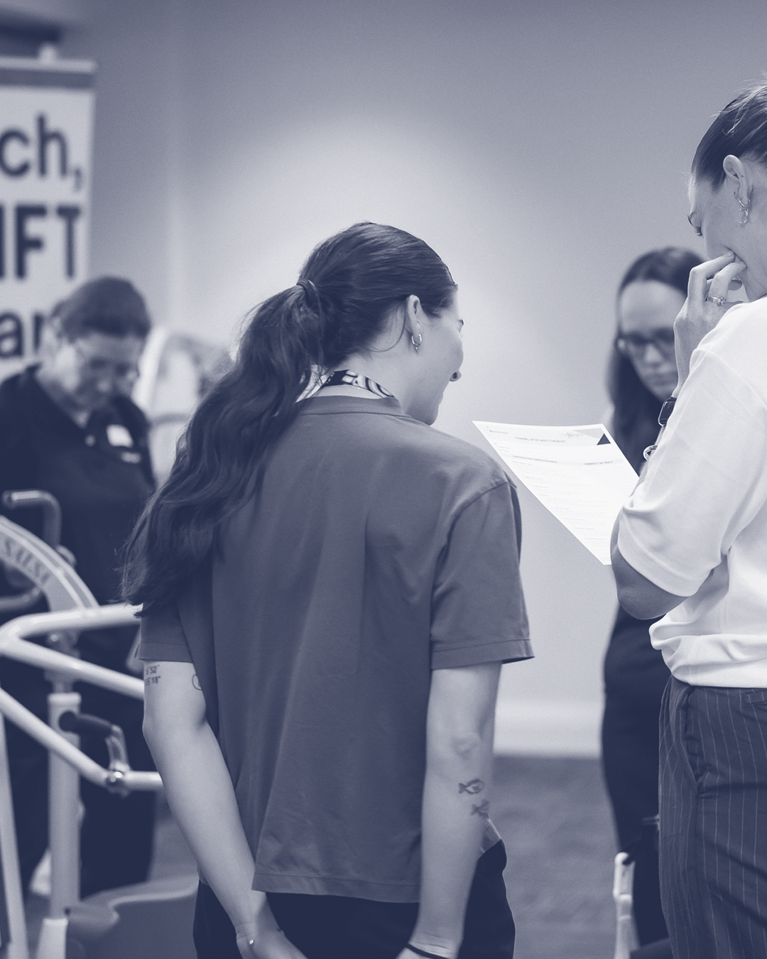 Group of people at a healthcare or vaccination site, with one woman in a uniform wearing a badge, engaging with a healthcare worker holding a sheet of paper, in a medical setting.
