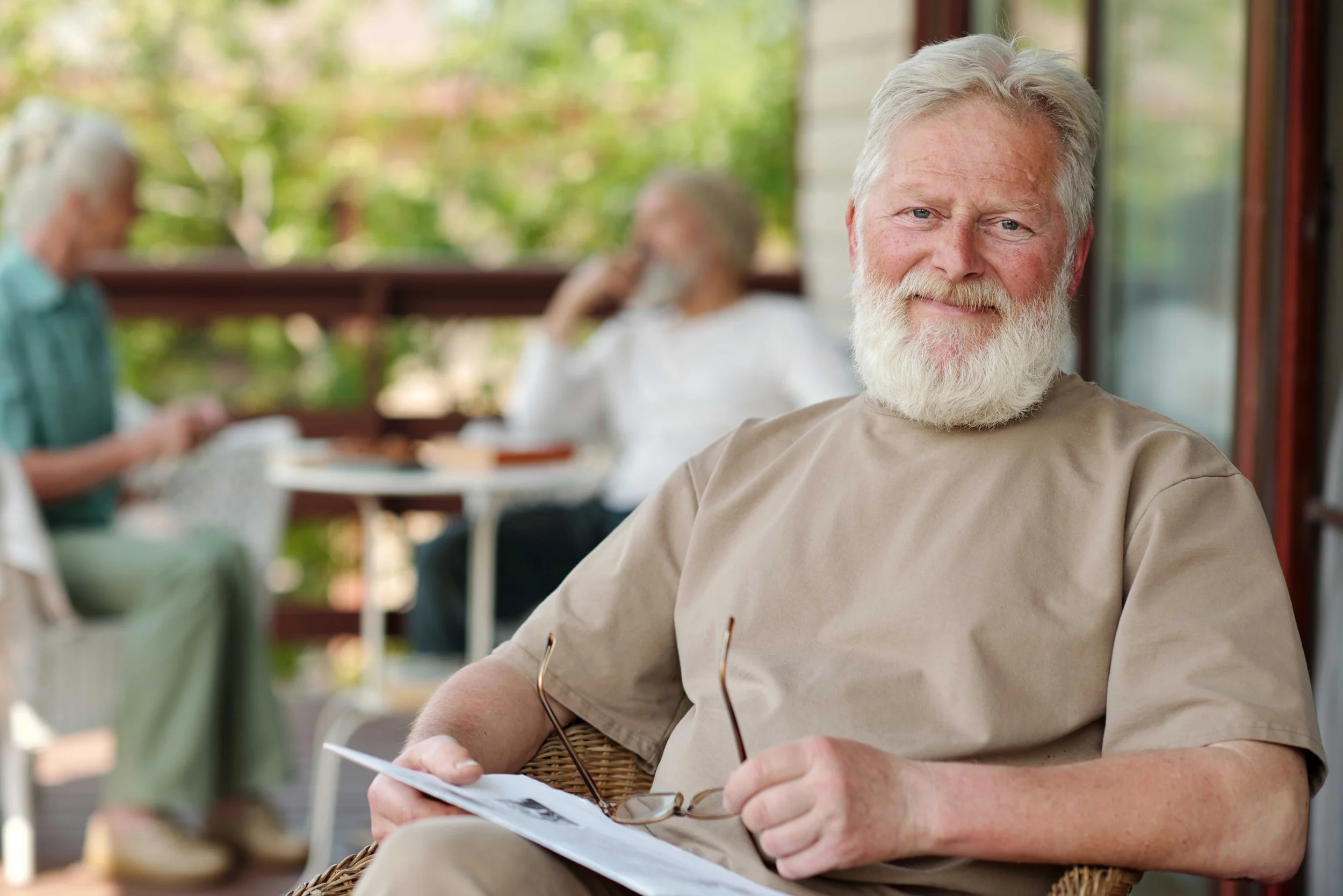 An elderly man with white hair and beard sitting outside on a patio, wearing a beige t-shirt, holding a pair of glasses and a paper, with a group of people blurred in the background.