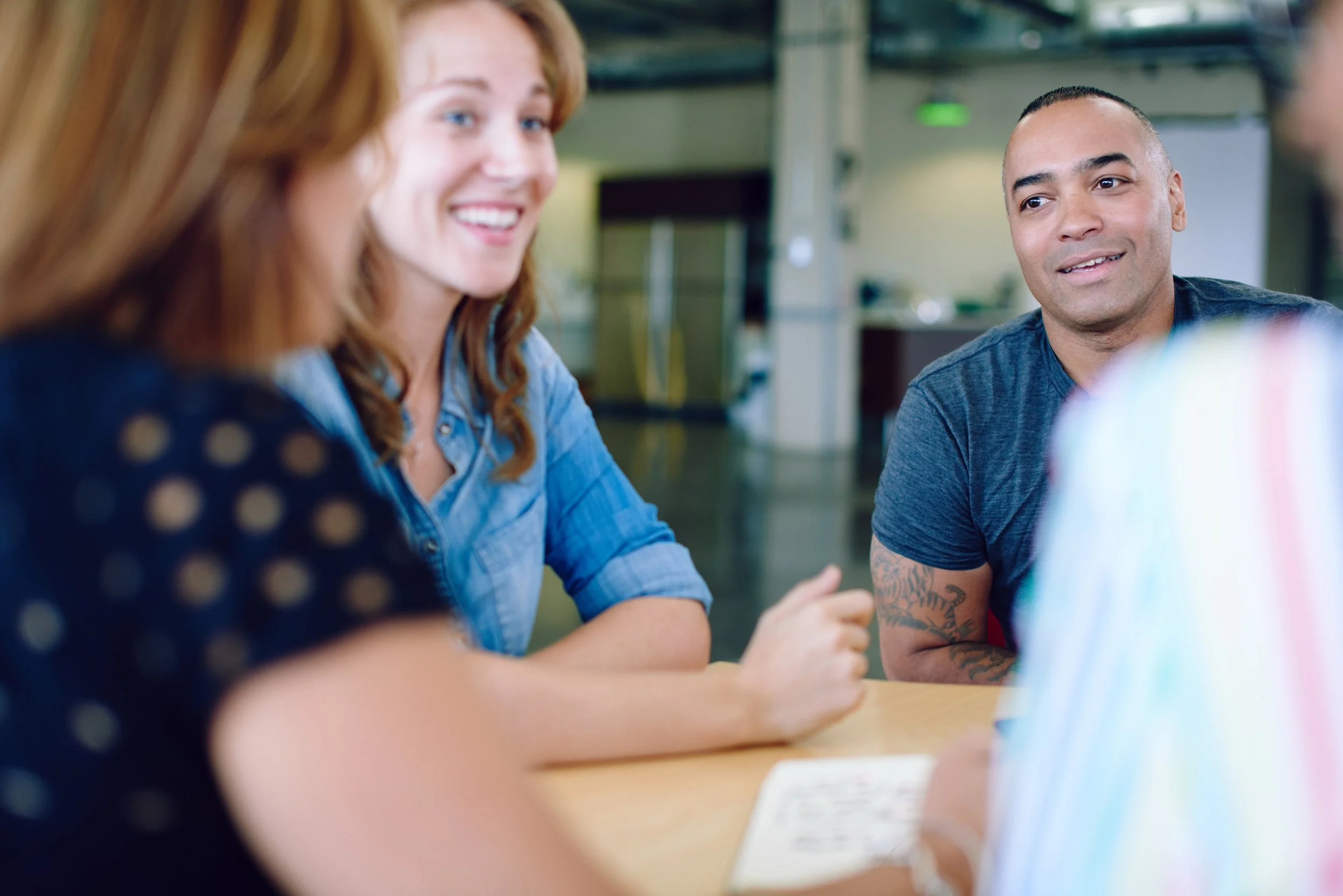 Four people sitting around a table engaged in a conversation, with focus on a woman with red hair smiling and a man with a tattoo on his arm listening attentively.