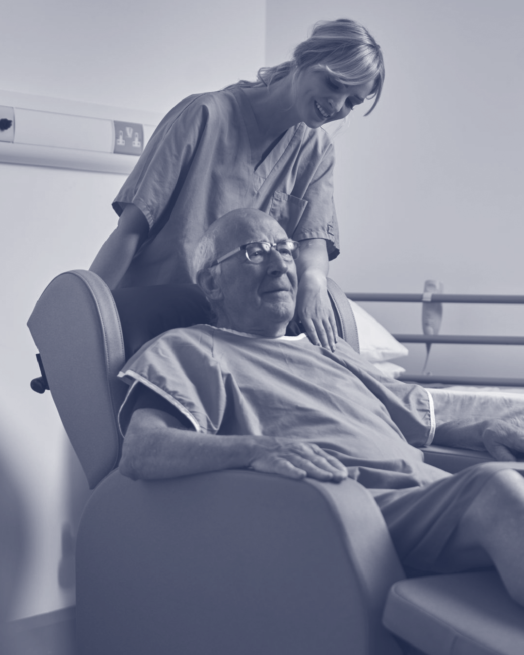 A nurse or caregiver smiles at an elderly man lying in a hospital bed, looking at him lovingly.