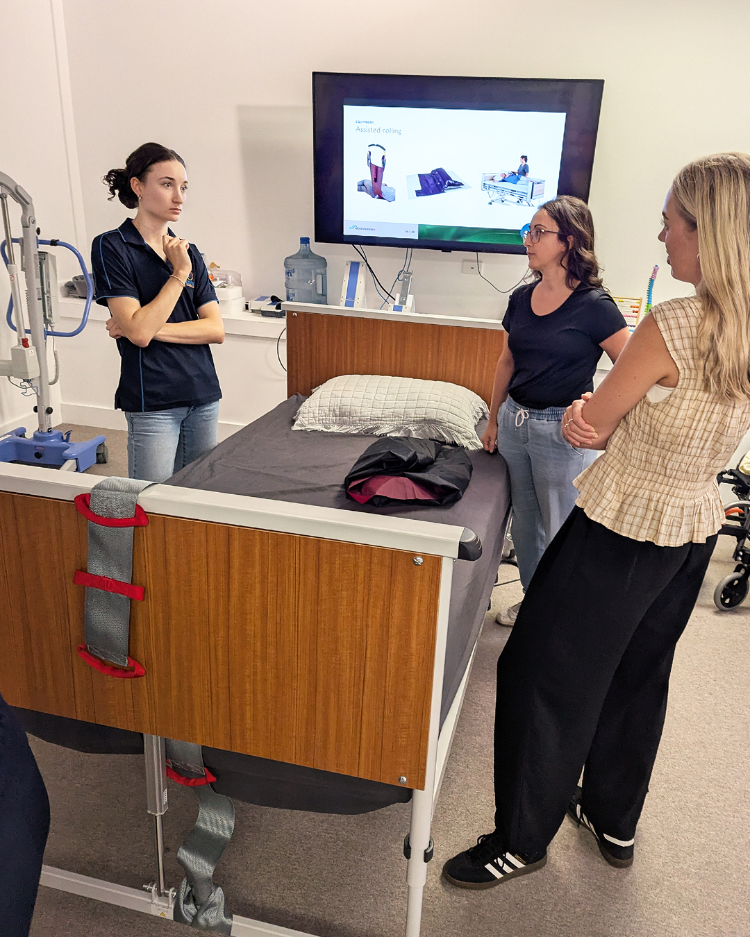 Medical training session in a hospital room with three women standing near a hospital bed, a large monitor displaying assistive rolling equipment, and medical supplies in the background.