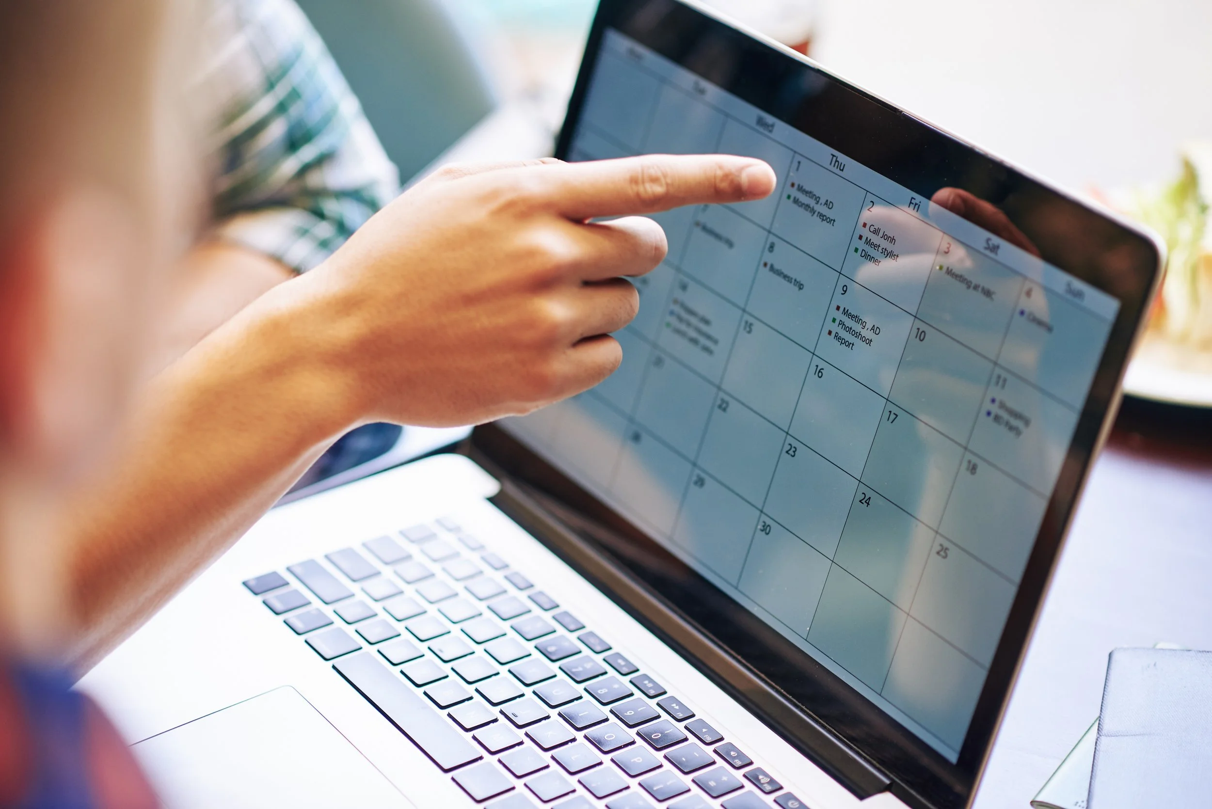 A person is using a tablet with a digital calendar app, while a laptop is open in front of them. The person is pointing at a date on the tablet.