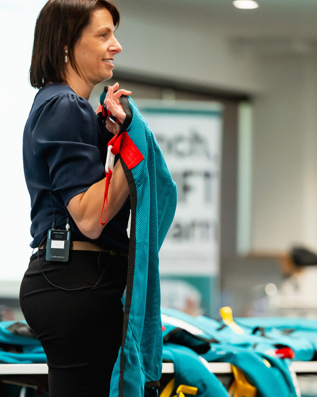 A woman with shoulder-length brown hair, wearing a dark blue blouse, holding a blue and red backpack, standing in front of a table with similar backpacks in an indoor setting.