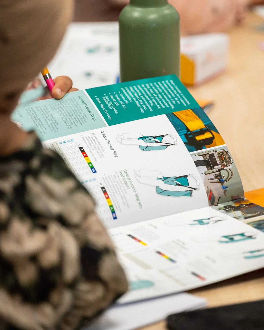 Person reading a catalog that features technical drawings and descriptions of career purpose sing clothing, with a green bottle and other objects on the table.