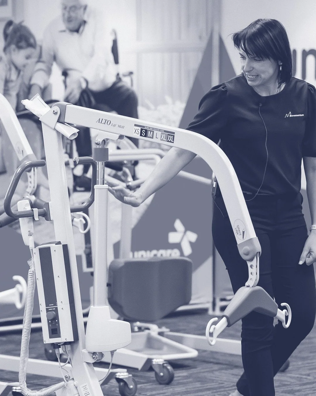 A woman in a black shirt using a medical robotic arm in a healthcare setting, with other people and medical equipment in the background.