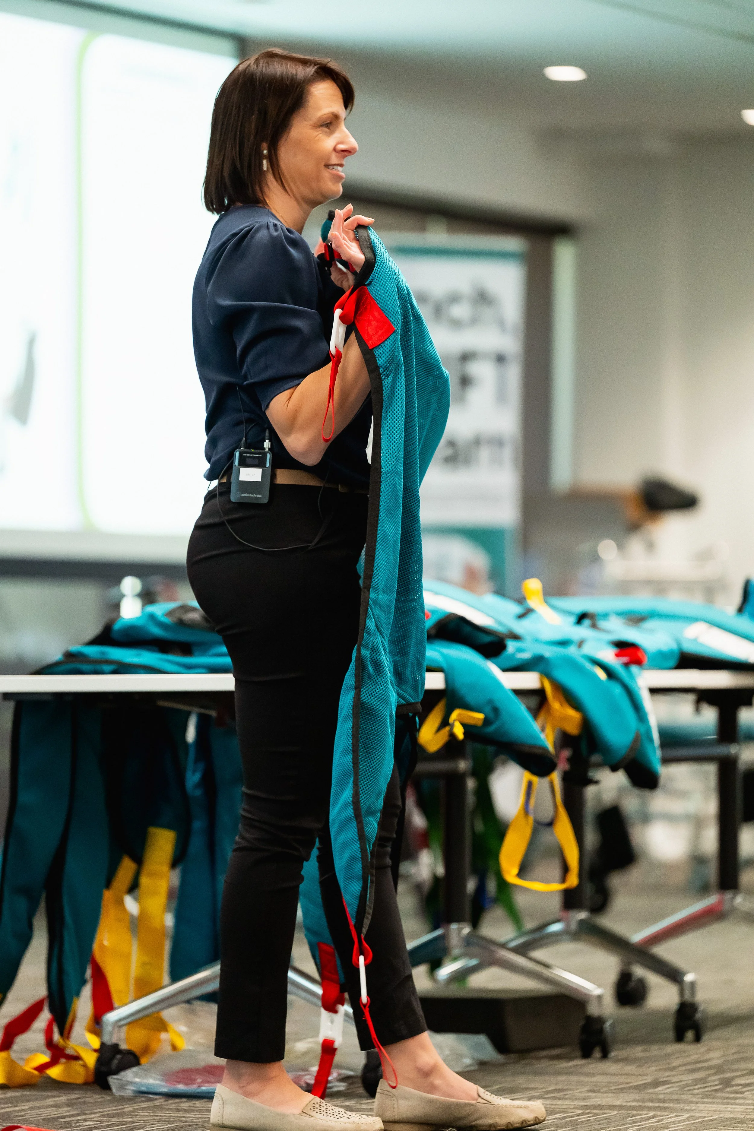 A woman in a navy blue shirt and black pants is holding a harness and preparing for a zipline activity at an indoor event space. There are colorful backpacks on a table behind her, and a stand with a poster in the background.